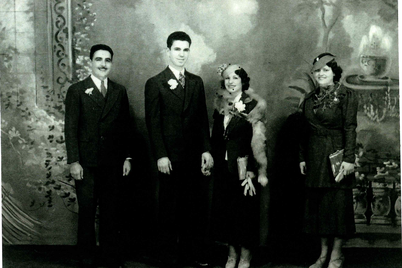 Black and white photograph of five people in vintage formal attire standing in front of a painted backdrop. Two men in suits and three women in dresses and hats, holding books or folders.