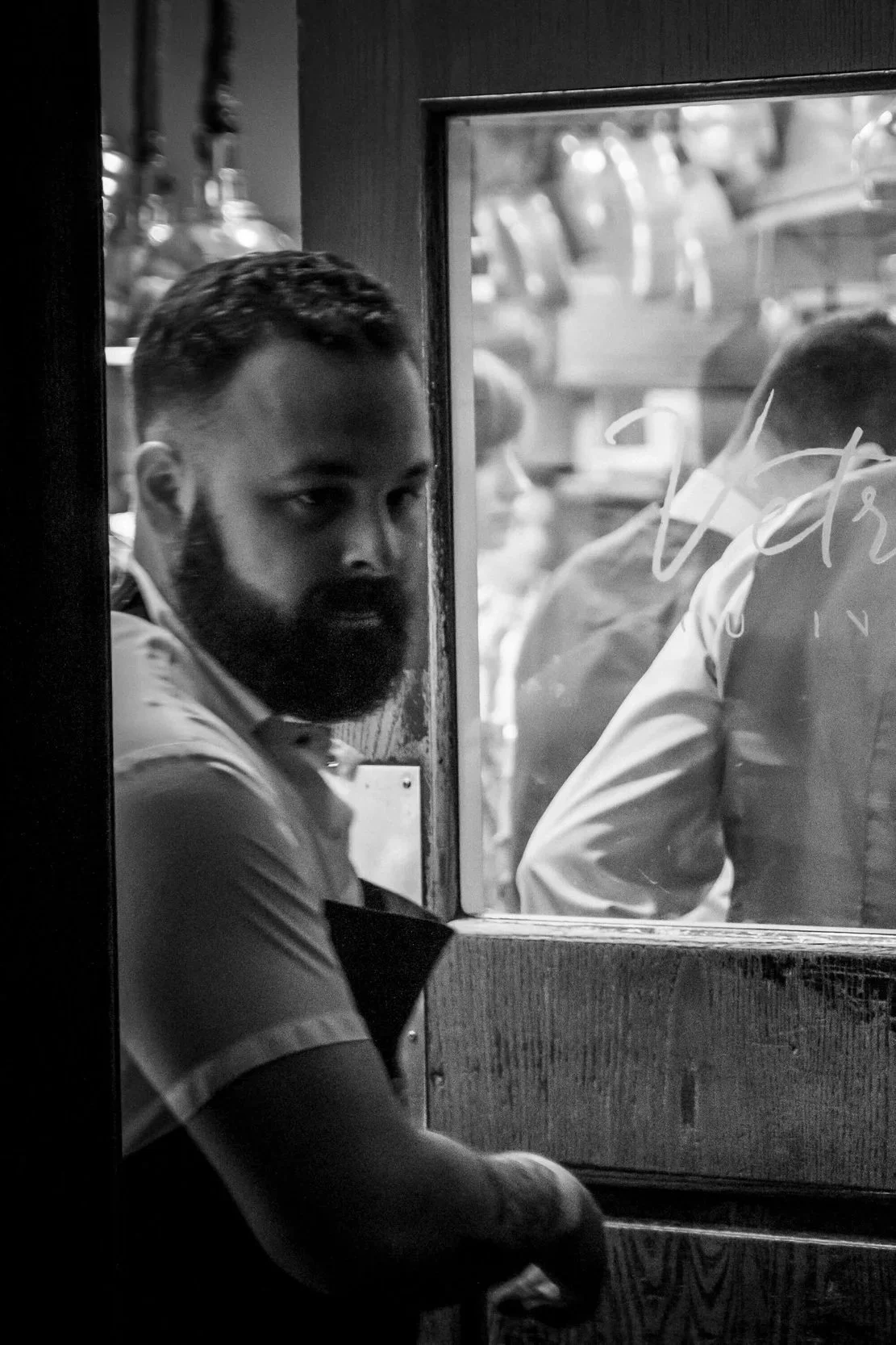 Black and white photo of a man with a beard and short hair sitting inside a restaurant or bar, looking through a window. The background outside the window shows a parking lot with several cars and people.