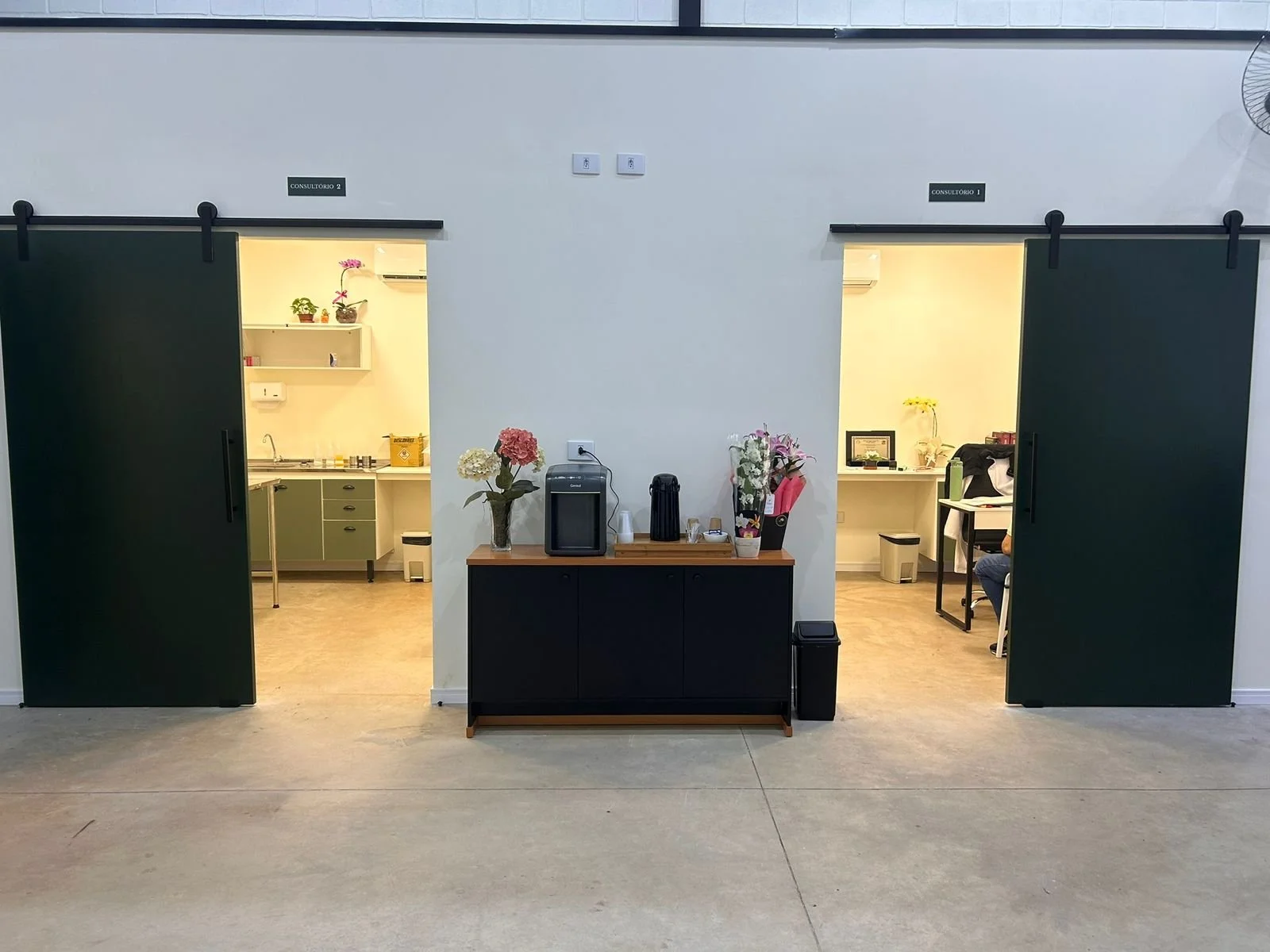 Salas de atendimento com portas de correr verdes, uma na esquerda e outra na direita, com sinais de consultórios 1 e 2 acima, e uma mesa no meio com flores e uma máquina de água ou café.