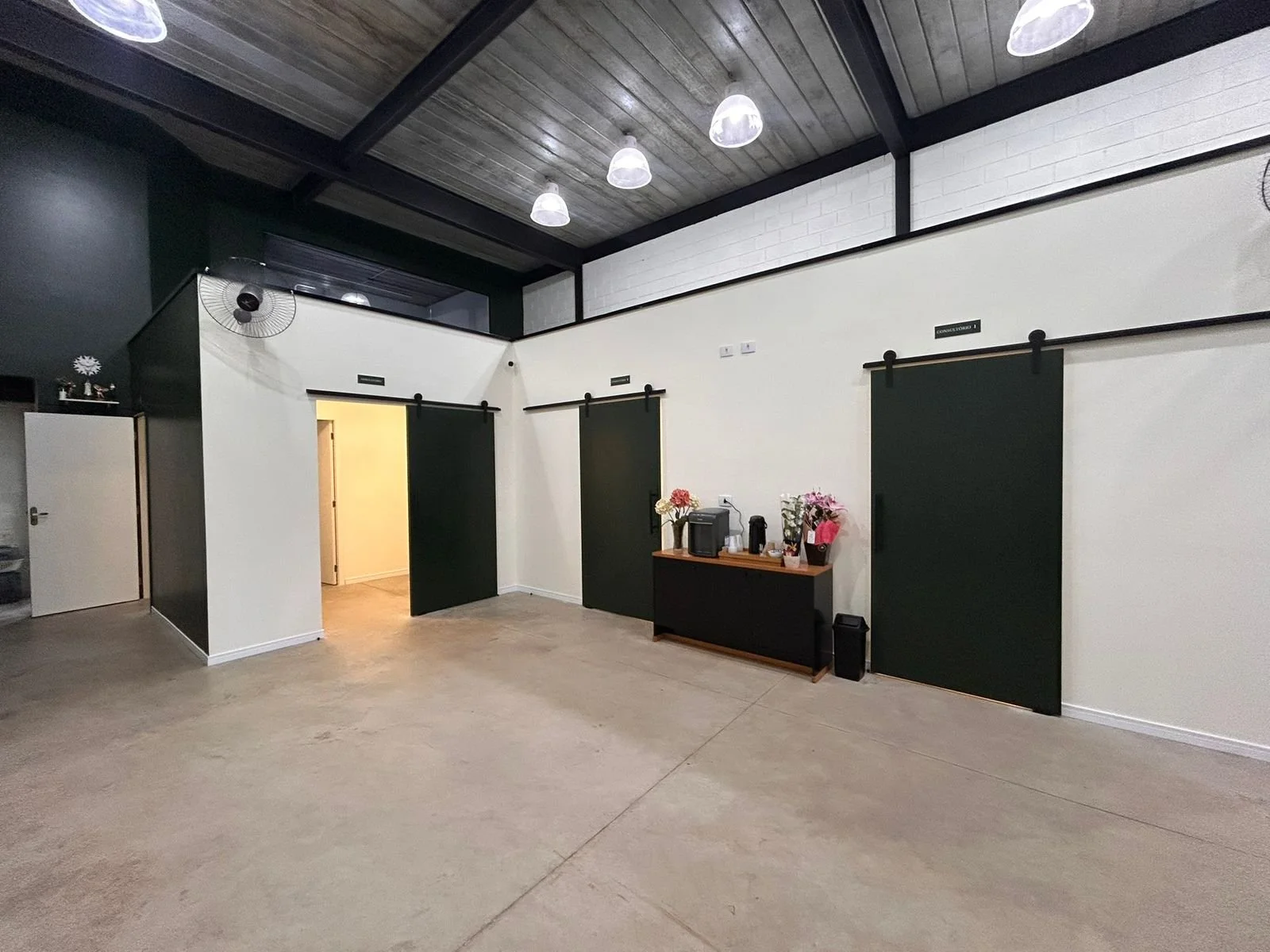 Sala com paredes brancas, teto de madeira com iluminação pendente, portas de correr verdes, balcão com flores, máquina de café e utensílios.