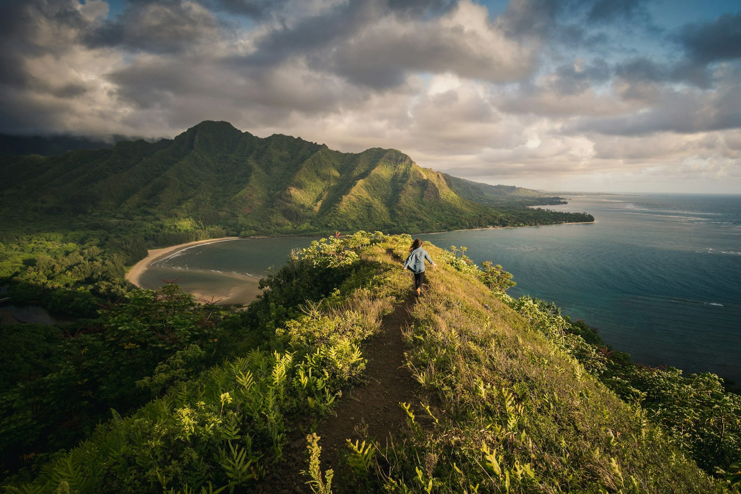 A person walking along a narrow trail on a lush green ridge overlooking a bay with a sandy beach, surrounded by mountains and ocean under a cloudy sky.