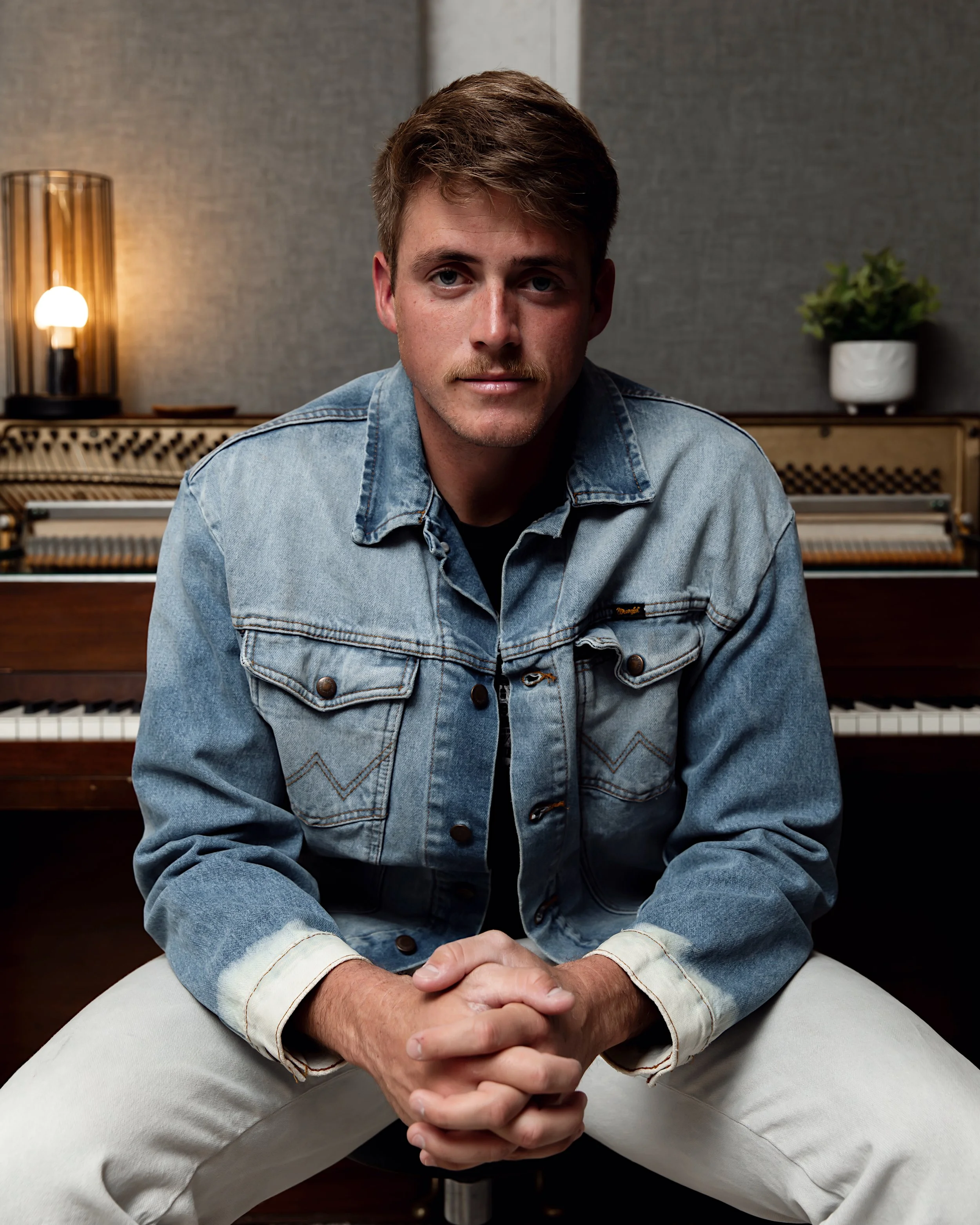 A young man wearing a denim jacket and white pants sitting in front of an upright piano with a lamp and potted plant in the background.