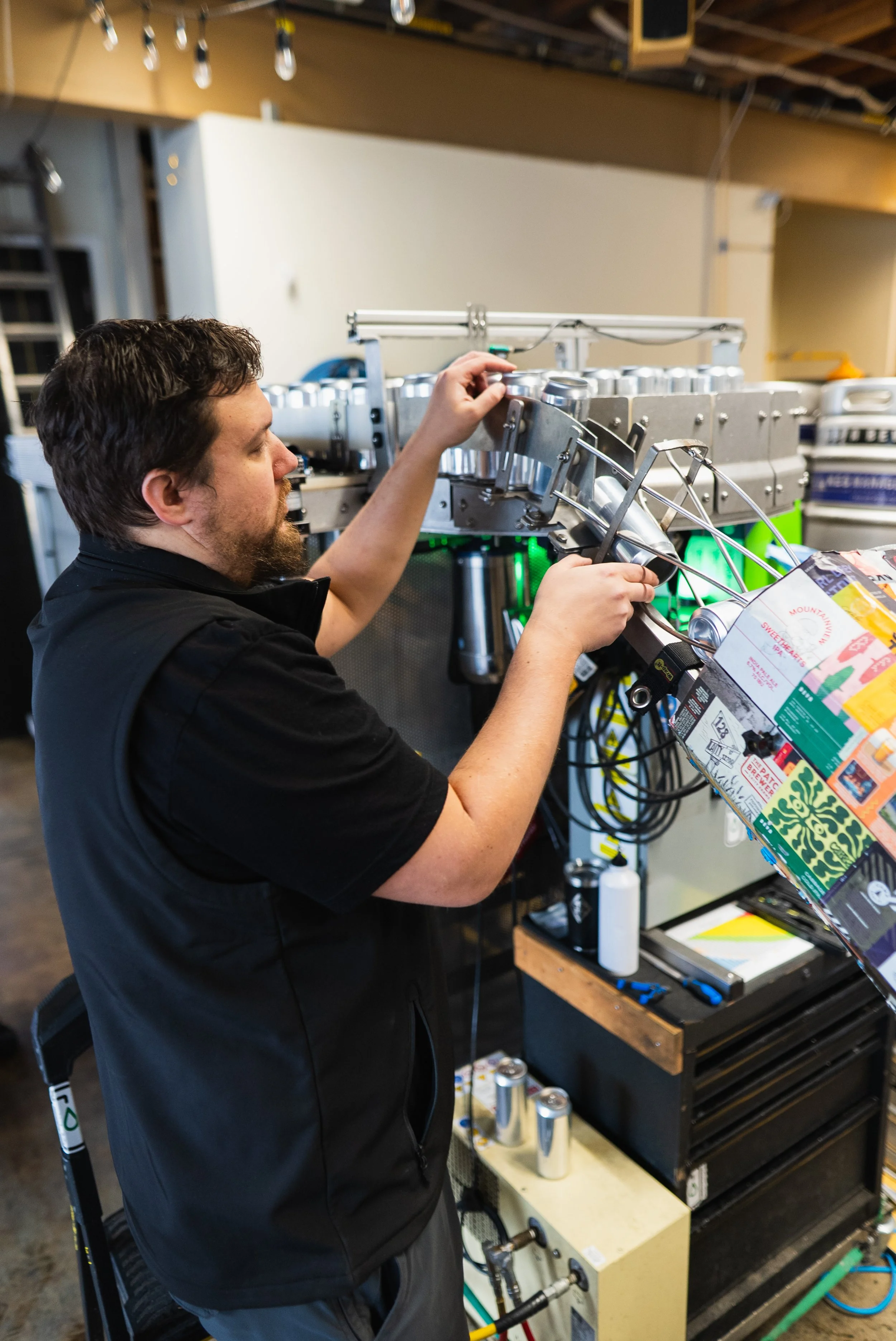 A man working on a machine assembling cans in a brewery or factory setting, with colorful packaging hanging nearby.