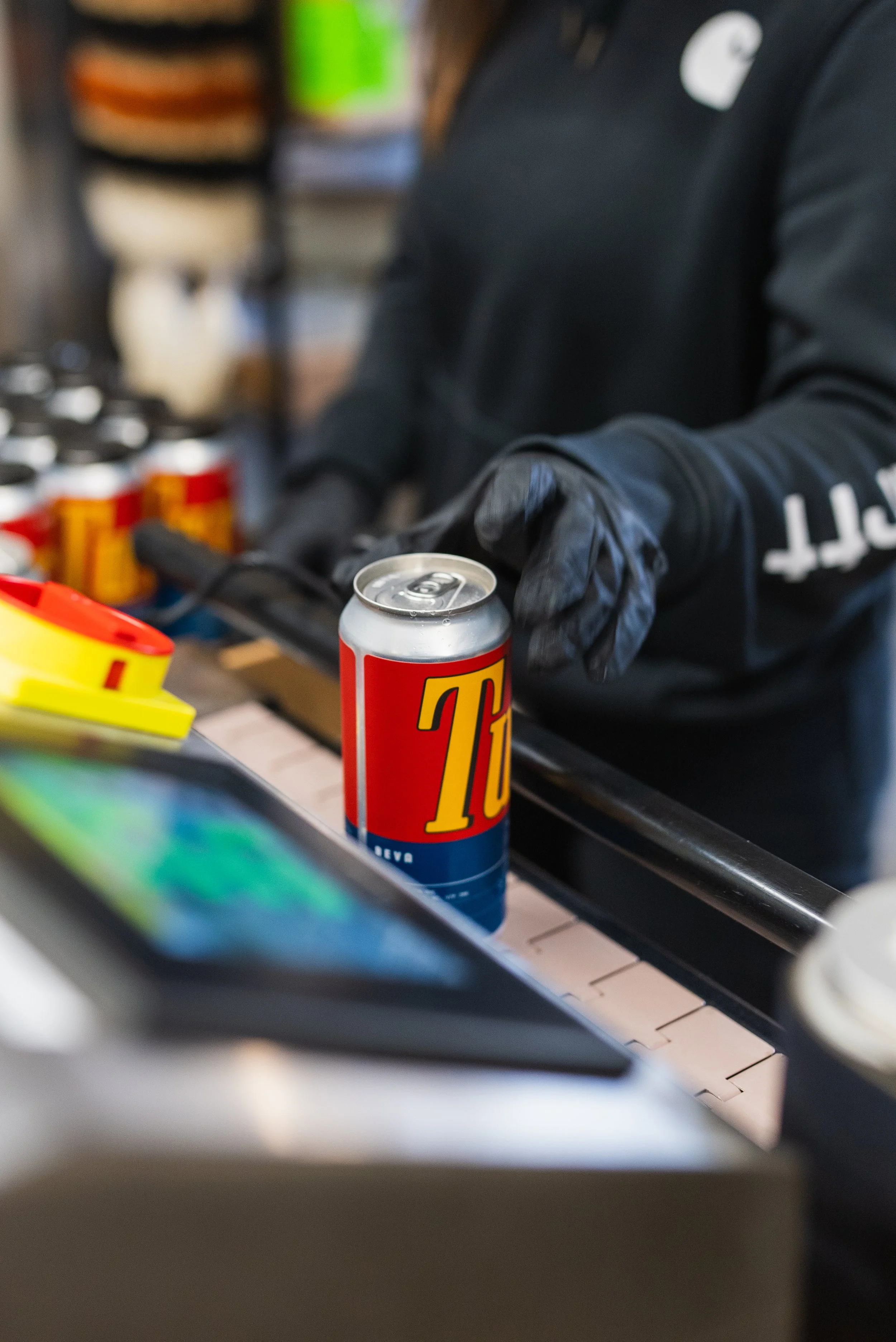 A person wearing black gloves and a black jacket is handling a can of Coors beer in a store or bar setting. Several other cans are visible in the background.