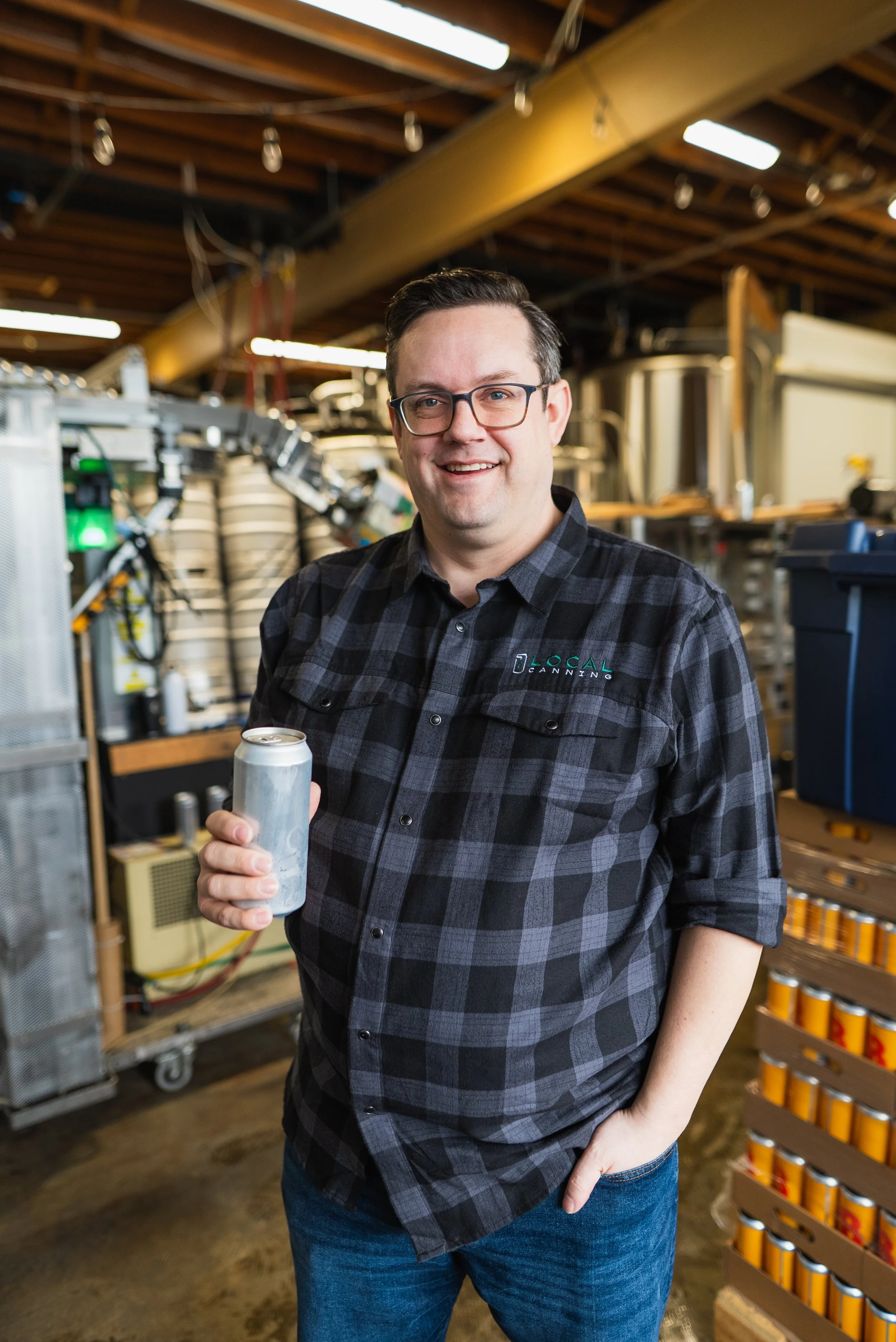 A man with glasses in a black plaid shirt holding a beverage can and smiling, standing in a brewery or taproom environment.