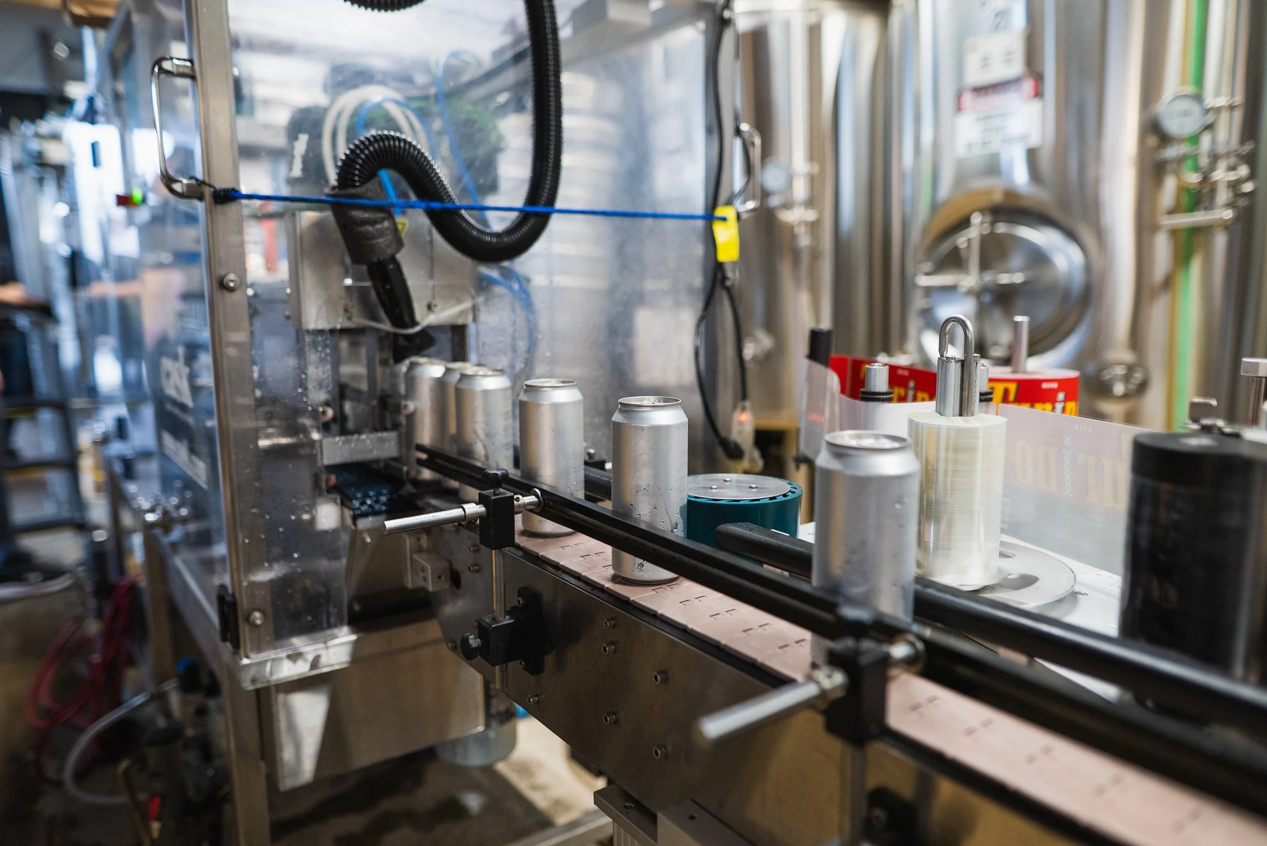 A close-up of a canning line in a brewery or factory, with silver cans moving along a conveyor, surrounded by industrial equipment and stainless steel tanks in the background.