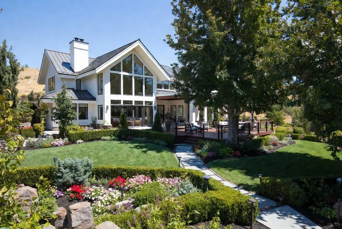 Luxurious Medford OR custom home: modern A-frame design, large windows, wooden deck, lush lawns, colorful flower beds, hedges & stone paths. Landscaping experts.