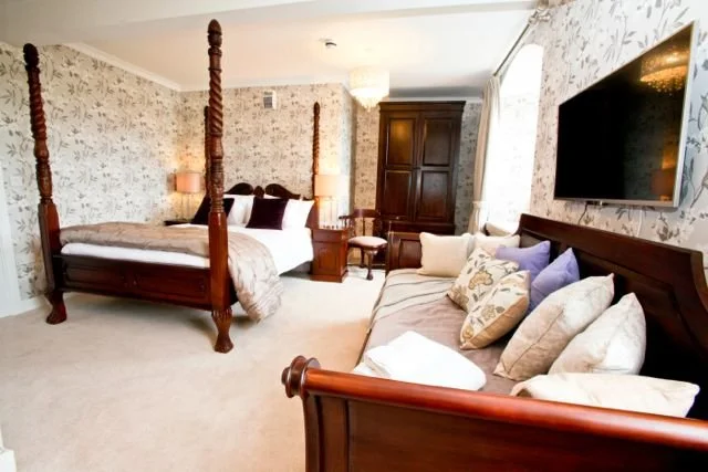 Bedroom with a four-poster bed, a sofa with multiple pillows, a large armoire, a flat-screen TV on the wall, and floral wallpaper.