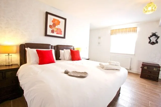 A bedroom with a large bed, white bedding, red pillows, and folded towels on the bed. There are two nightstands with lamps, a framed flower art piece on the wall, and a window with a beige blind.