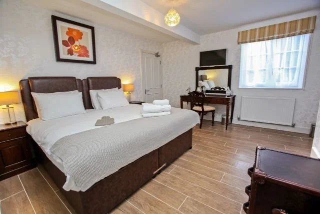 Hotel room with a large bed, nightstands with lamps, a framed floral artwork on the wall, a window with curtains, a mirror, and a small desk with chair.