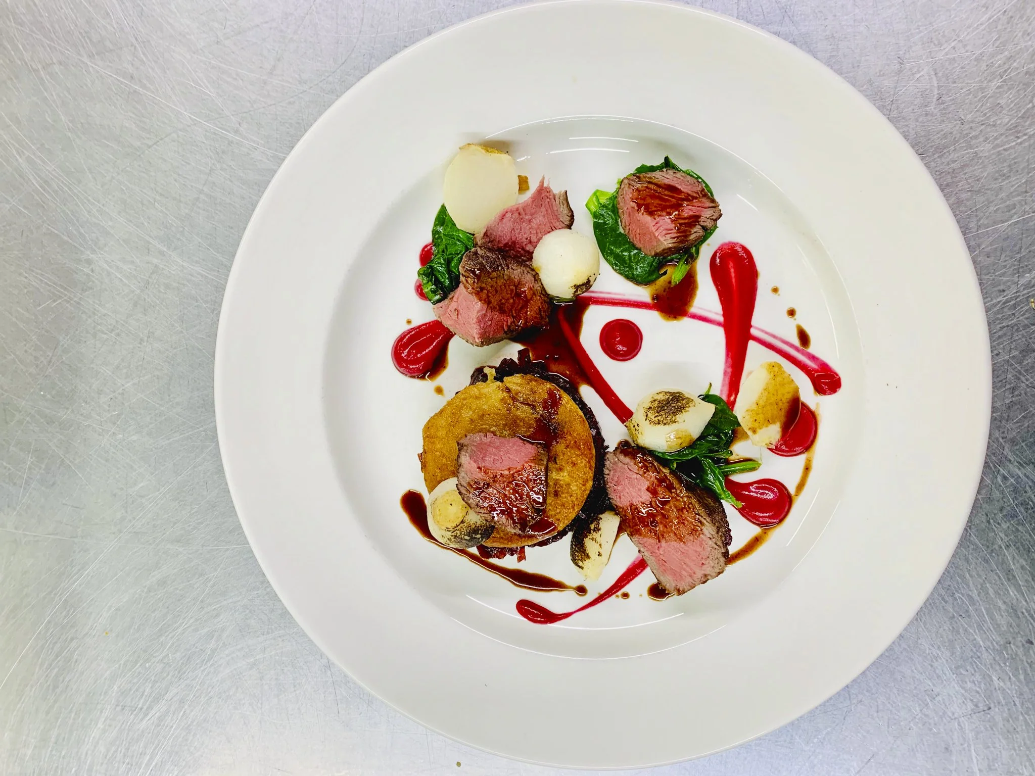 A plated gourmet dish with slices of medium-rare steak, mashed potatoes, vegetables, and artistic red sauce drizzles.