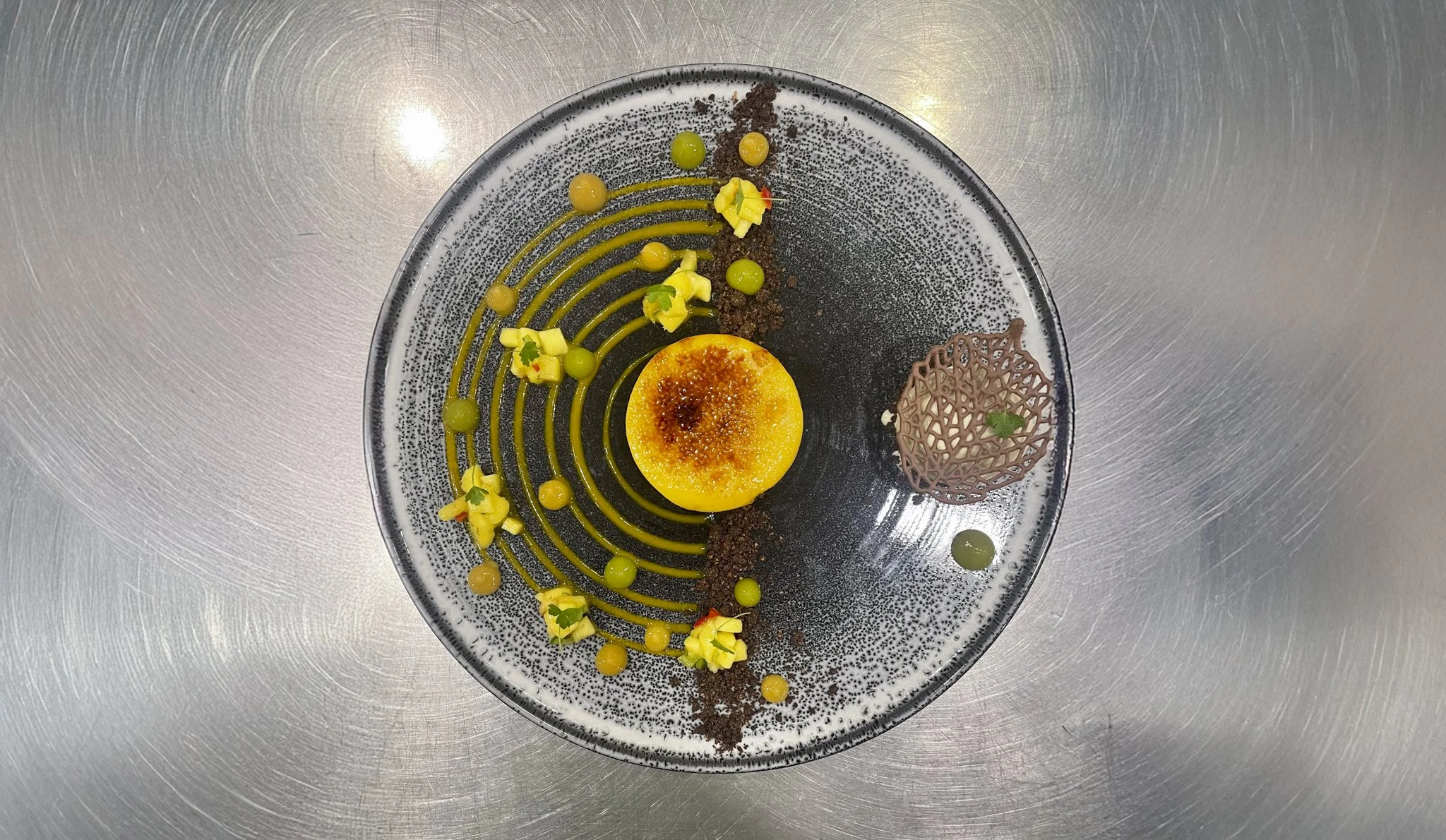 Elegant dessert plate with a caramelized tart in the center, surrounded by yellow and green sauce dots, yellow flower-shaped decorations, a chocolate lattice garnish, and a dusting of chocolate crumbs on a speckled black and white ceramic plate.
