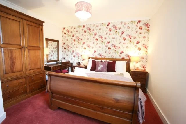 Bedroom with a wooden bed, floral wallpapered accent wall, wooden wardrobe, nightstands with lamps, and a pink carpet.