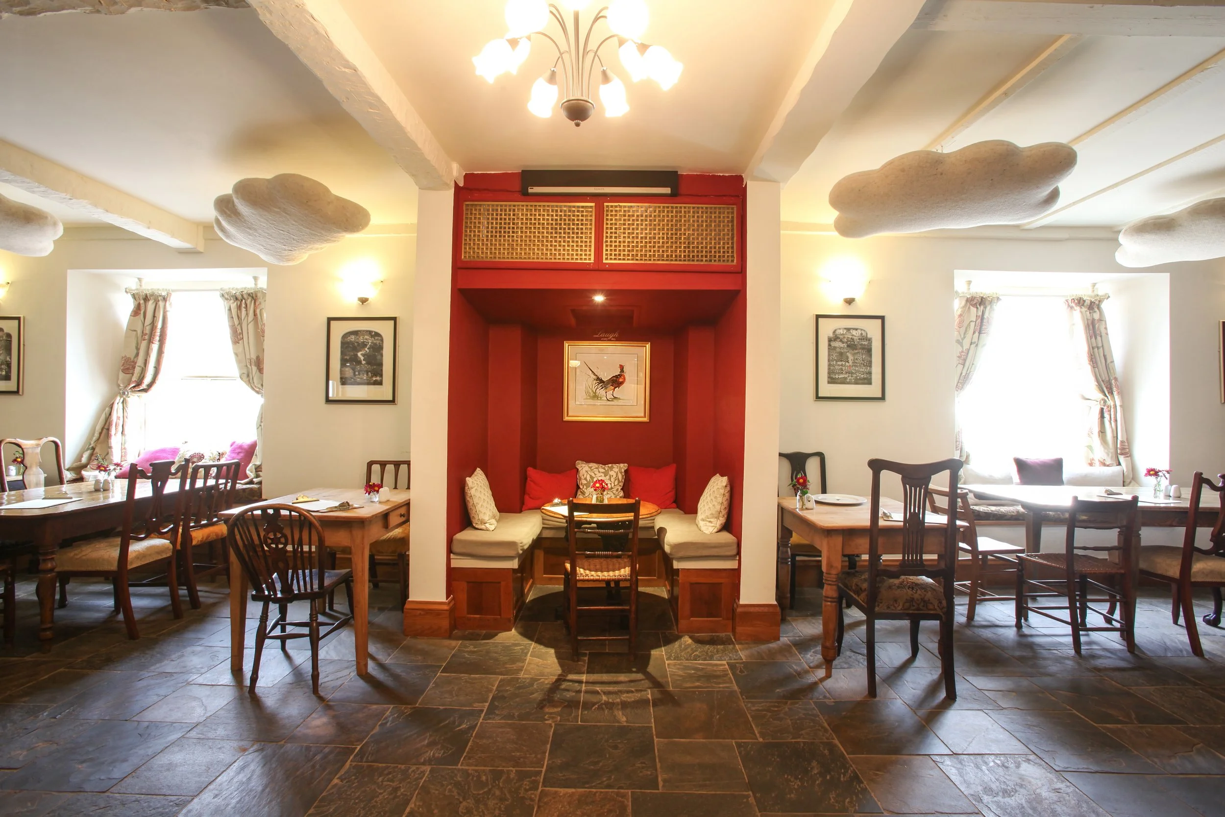 Interior of a cozy restaurant with wooden tables and chairs, decorative curtains, framed pictures on the walls, cloud-shaped ceiling fixtures, and a red alcove in the center with cushions and a framed picture of a rooster.