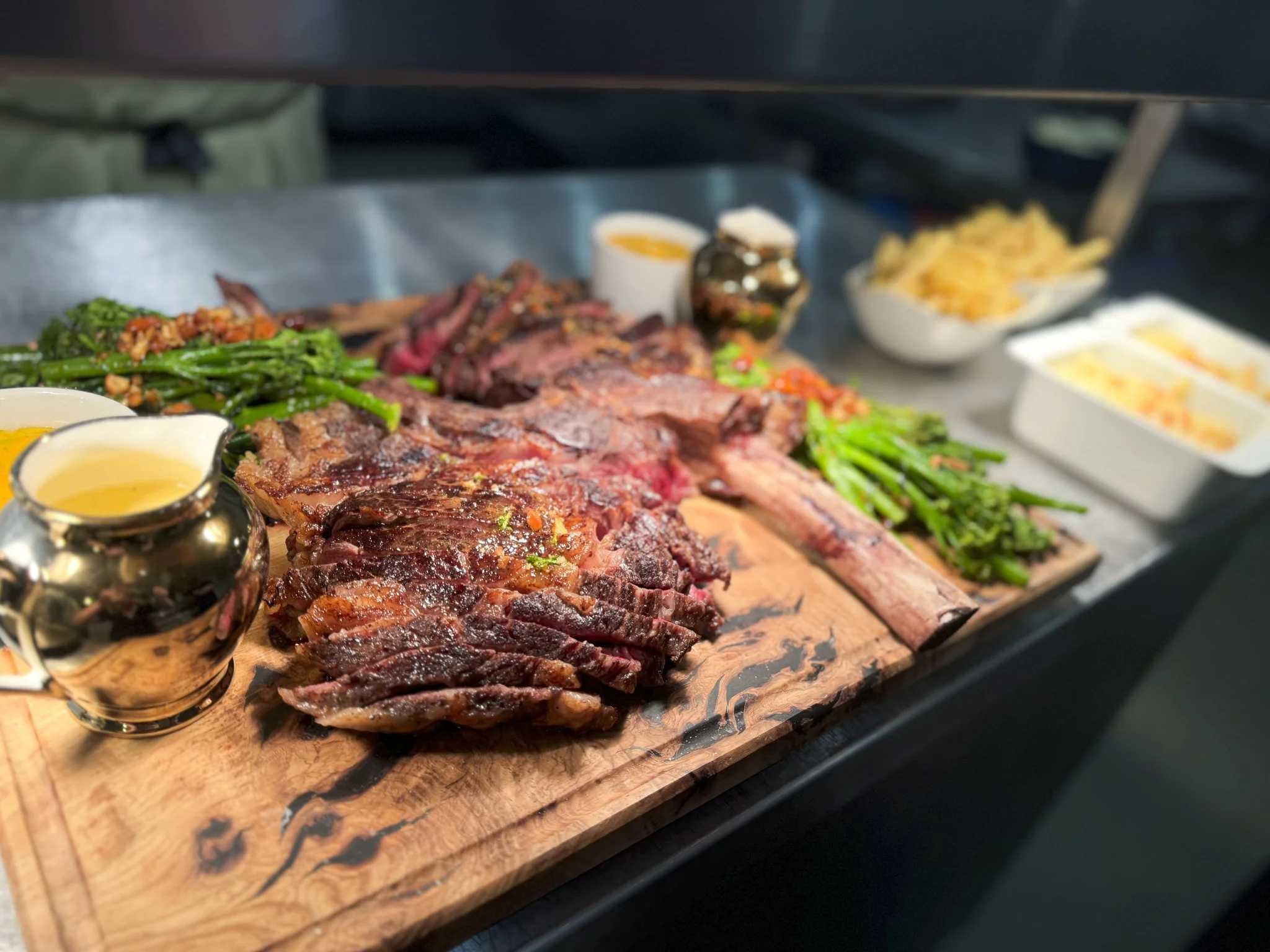 A wooden serving board with sliced cooked steak, green beans, and various condiments and sauces, with bowls of cheese and toppings in the background.