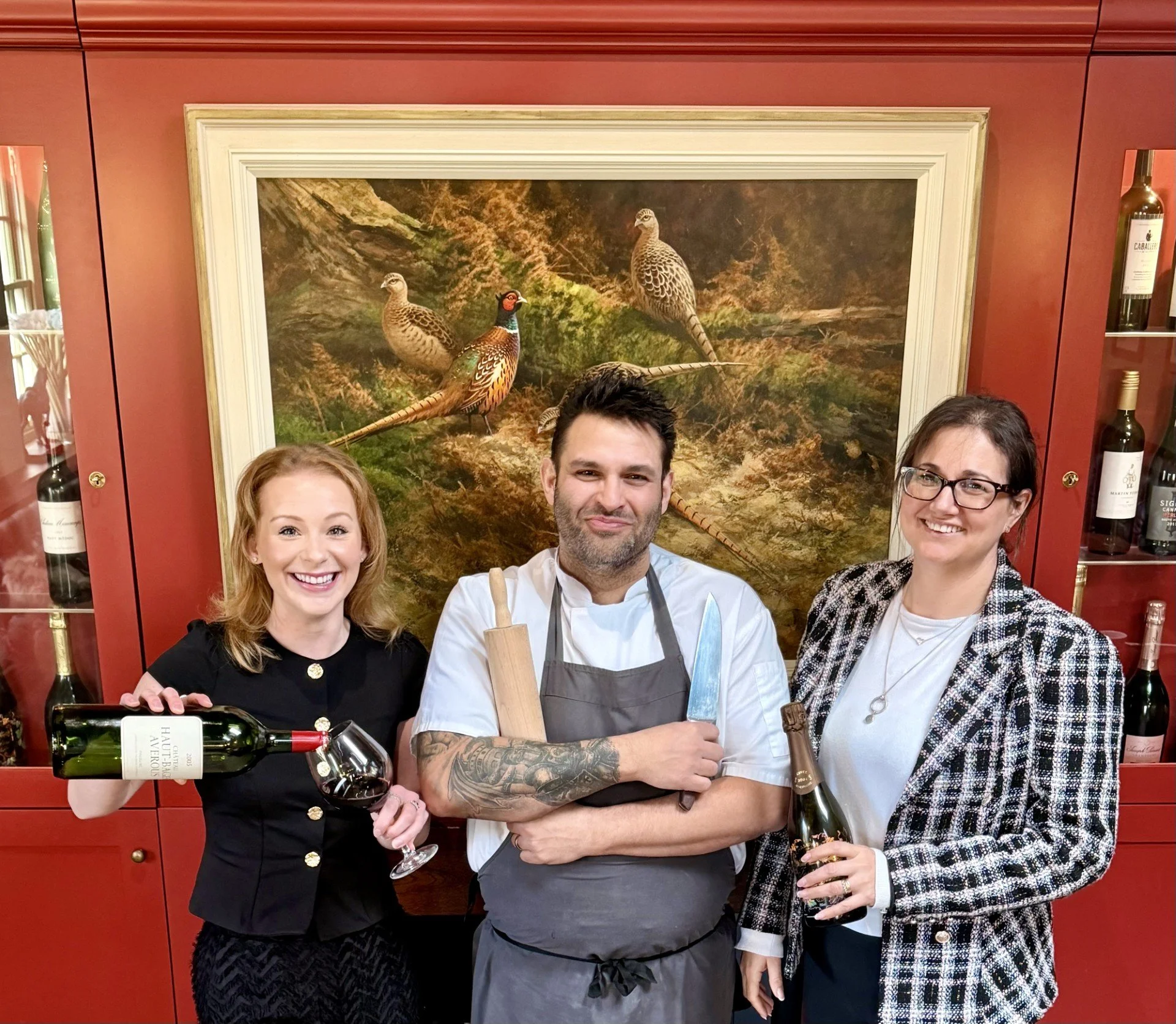 Three people standing in a restaurant, smiling at the camera. The woman on the left is pouring wine from a bottle, the man in the middle is holding a knife and rolling pin, and the woman on the right is holding a bottle of champagne. Behind them is a