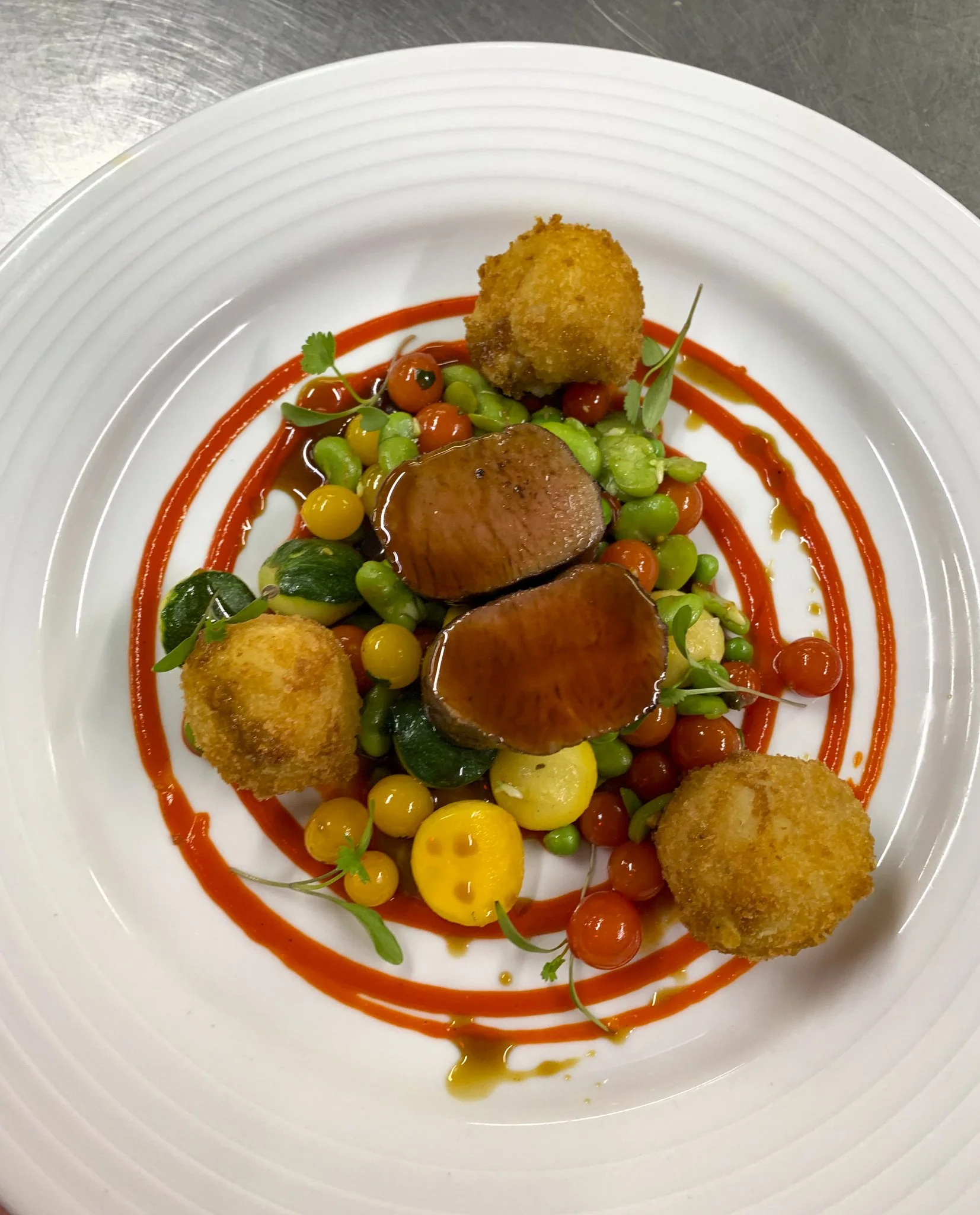 A gourmet dish featuring two slices of glazed meat, surrounded by colorful vegetables and garnished with microgreens, served with a decorative drizzle of red sauce on a white plate.