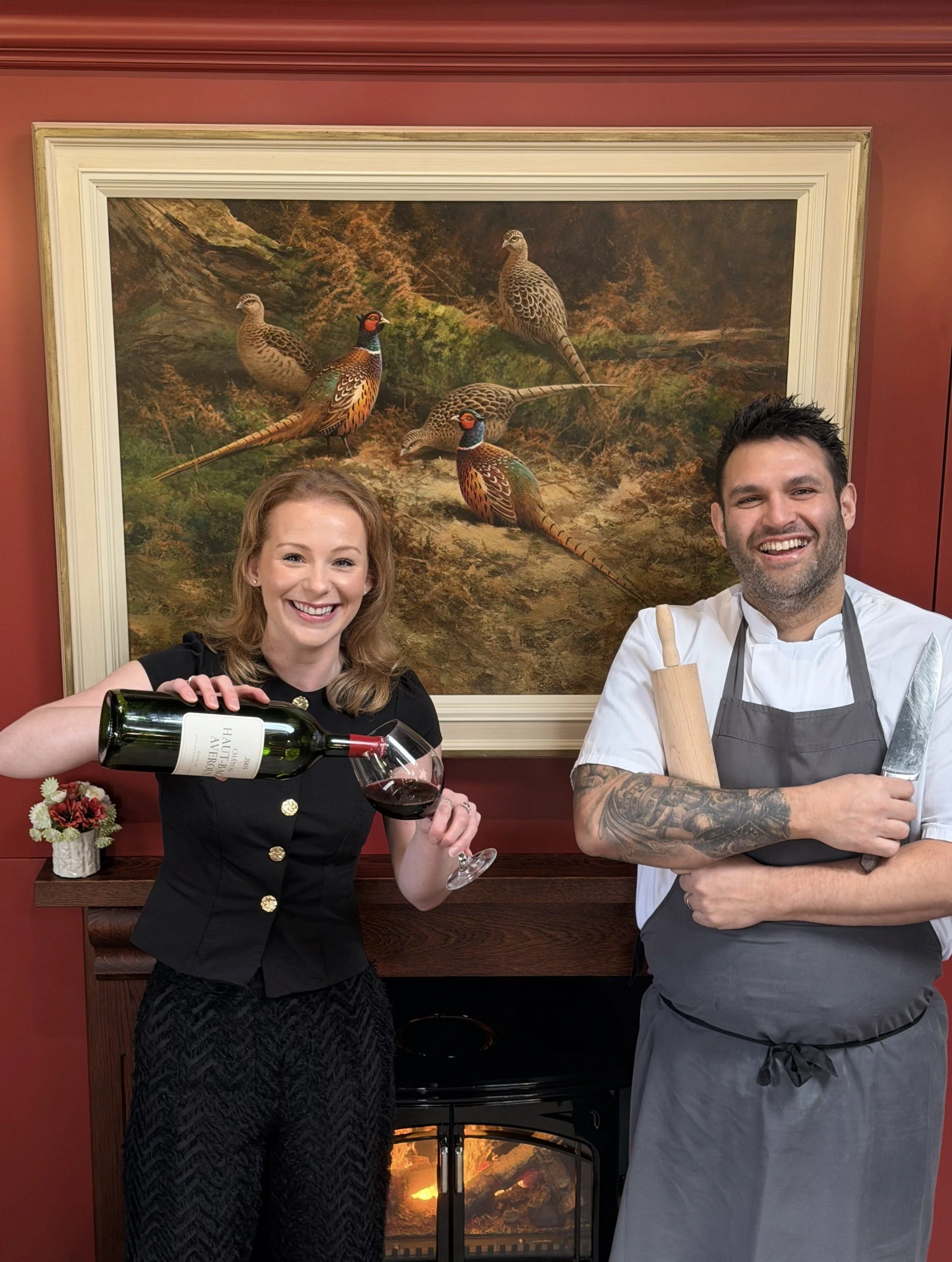 A woman in black holding a glass of red wine and a man in a chef's outfit with a rolling pin and knife, standing in front of a painting of birds and a fireplace.