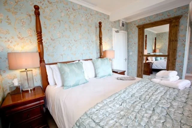 A bedroom with a four-poster bed, light blue pillows, beige wallpaper with floral patterns, two bedside lamps, a mirror showing another bed, and neatly folded towels on the bed.