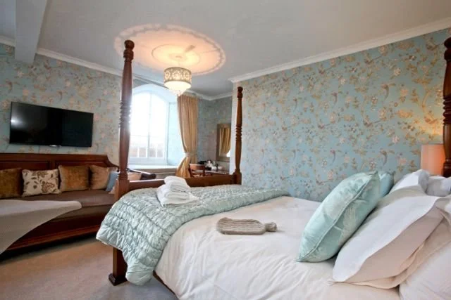 A bedroom with a four-poster bed, neatly made with light-colored bedding and pillows. There is a sofa with pillows along the wall, a window with curtains, and a wall-mounted flat-screen TV. The room features floral wallpaper and a ceiling light fixture.