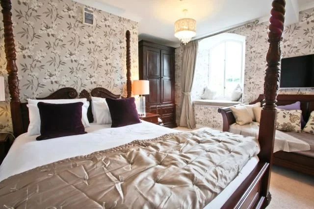 A bedroom with a four-poster bed, floral wallpaper, a window with curtains, a wooden armoire, a sofa with pillows, and a flat-screen TV