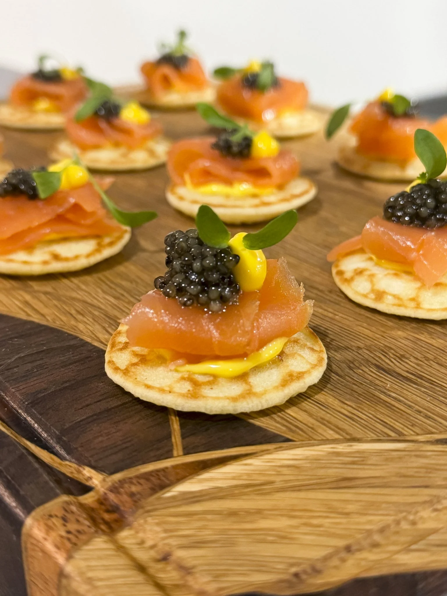 Smoked salmon canapé with caviar, microgreens, and yellow sauce on a small toasted bread base, arranged on a wooden serving tray.