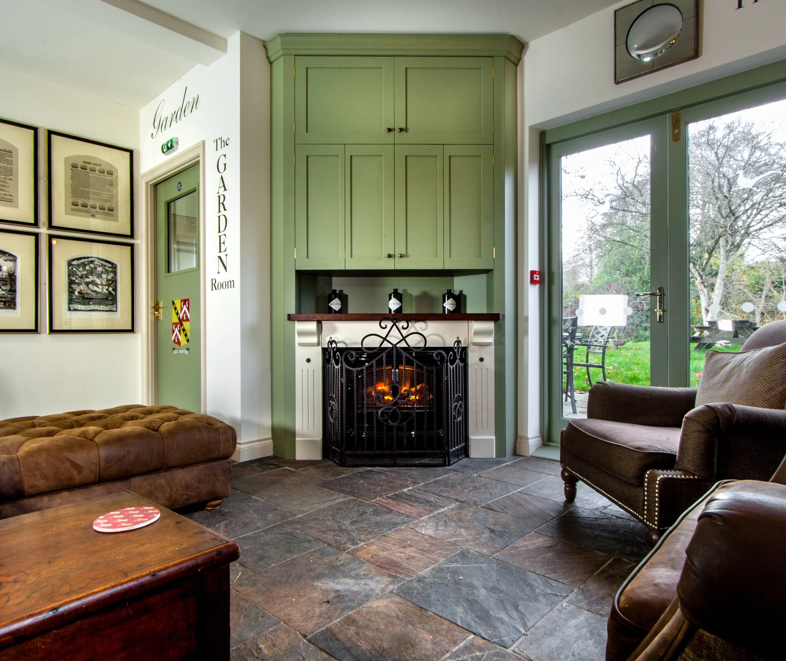 Cozy living room with stone tile floor, brown leather sofa, and a green corner cabinet around a fireplace with a decorative iron screen. A glass door leads outside to a garden with patio furniture.
