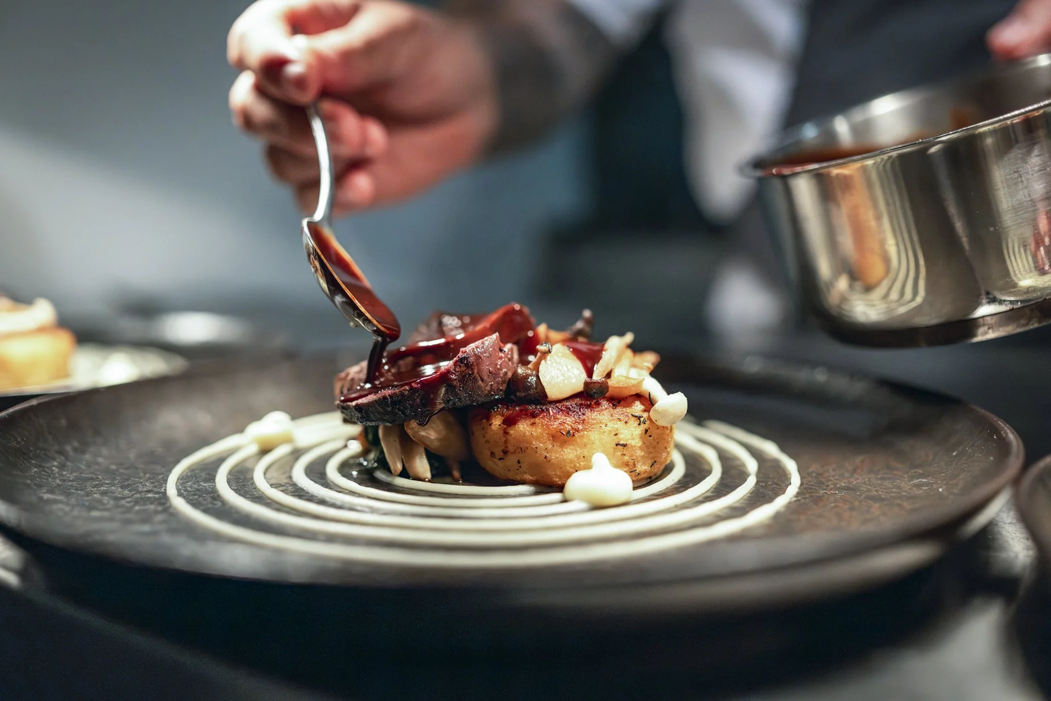 Chef drizzling sauce over a plated gourmet dish with a seared scallop, mushrooms, and white sauce decorative swirl on a dark textured plate.
