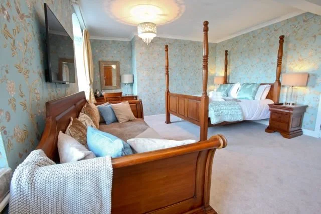 A bedroom with a wooden four-poster bed, a sofa with pillows, a wall-mounted TV, a chandelier, and floral wallpaper.