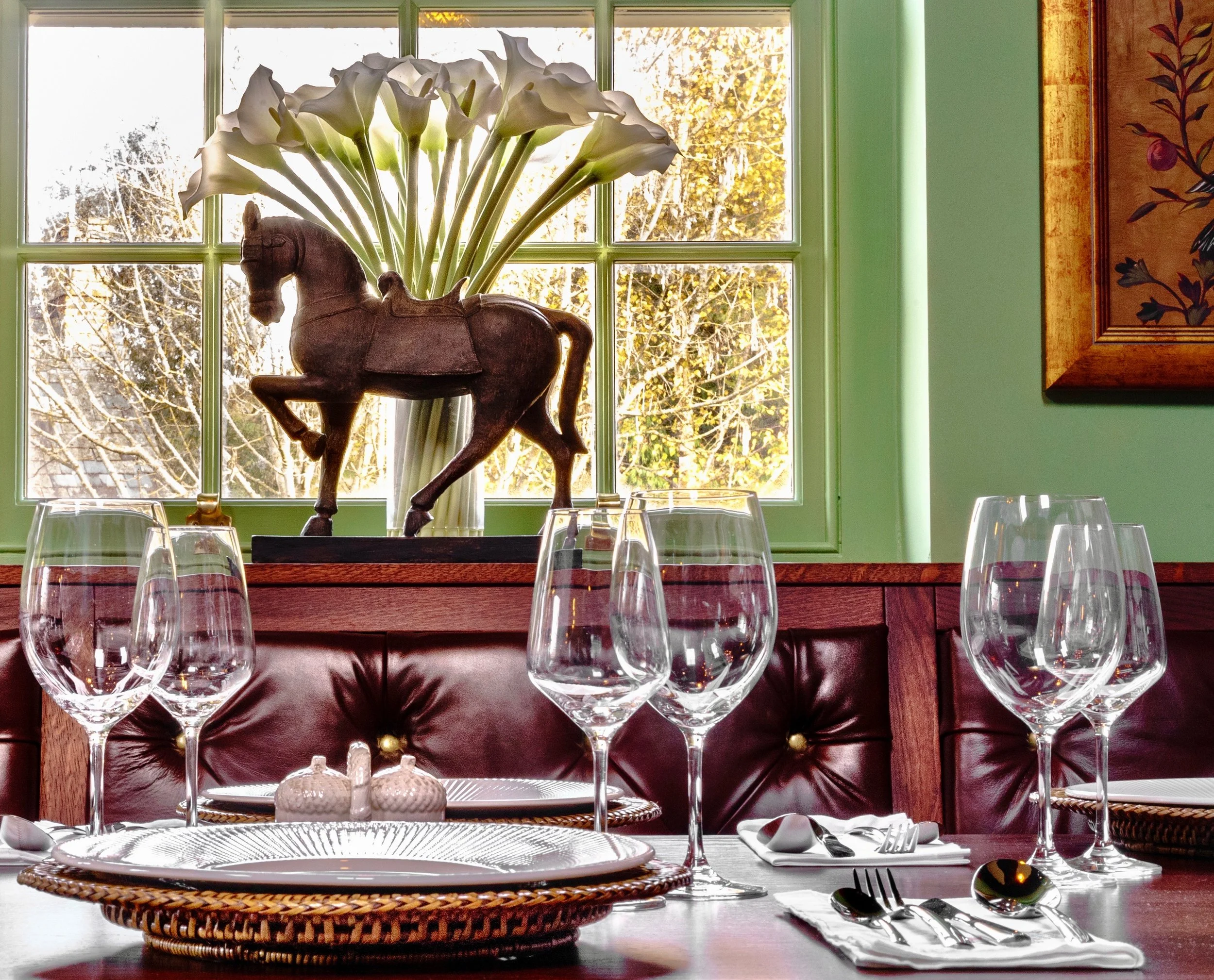Dining table set with wine glasses, plates, and cutlery, with a vases of white calla lilies and a decorative wooden horse sculpture on the windowsill in front of a window with a view of trees outside.