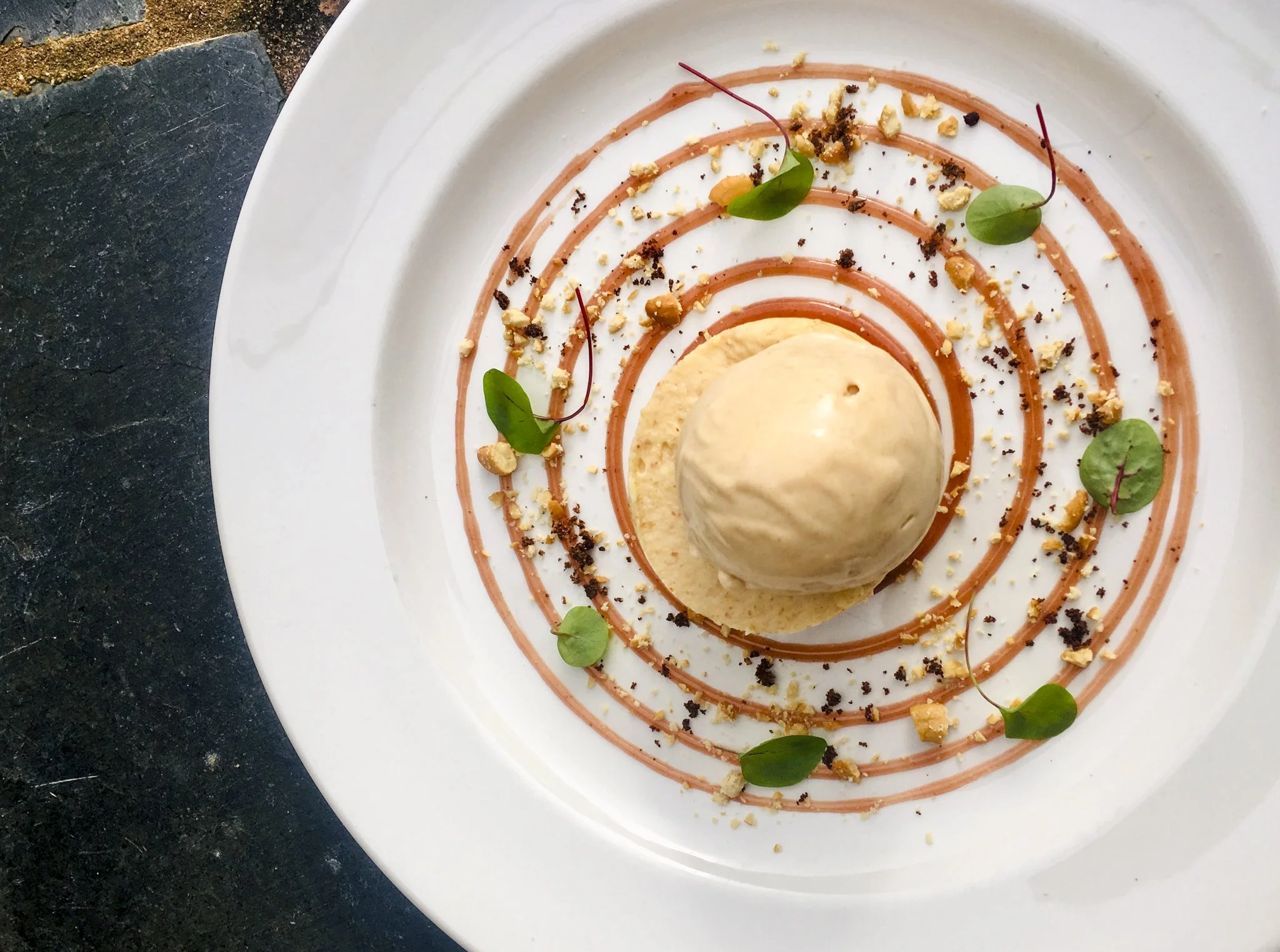 Vanilla ice cream scoop on a plate decorated with caramel, chocolate crumbs, chopped nuts, and microgreens, with a circular pattern of caramel drizzle.