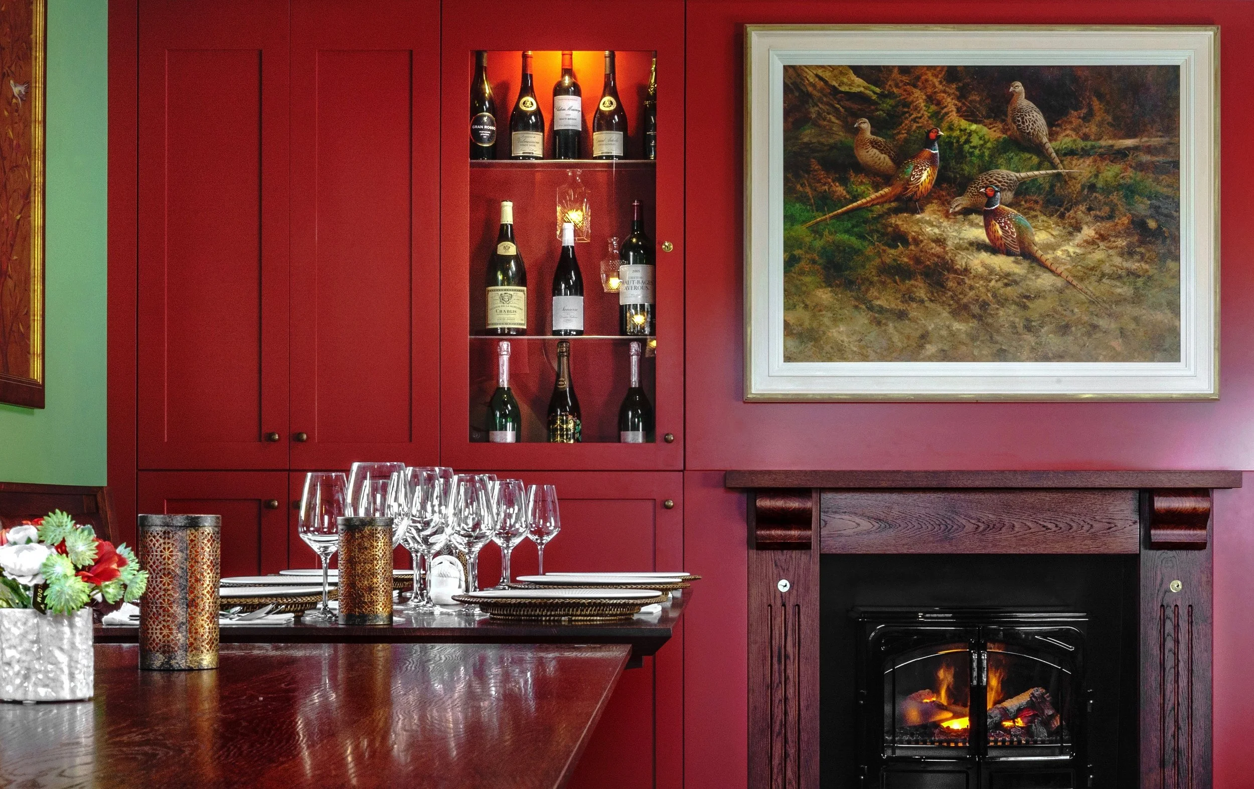 A dining room with a red wall, a fireplace, a framed painting of birds, a table set with wine glasses, candles, and a floral centerpiece, and a built-in cabinet with wine bottles.