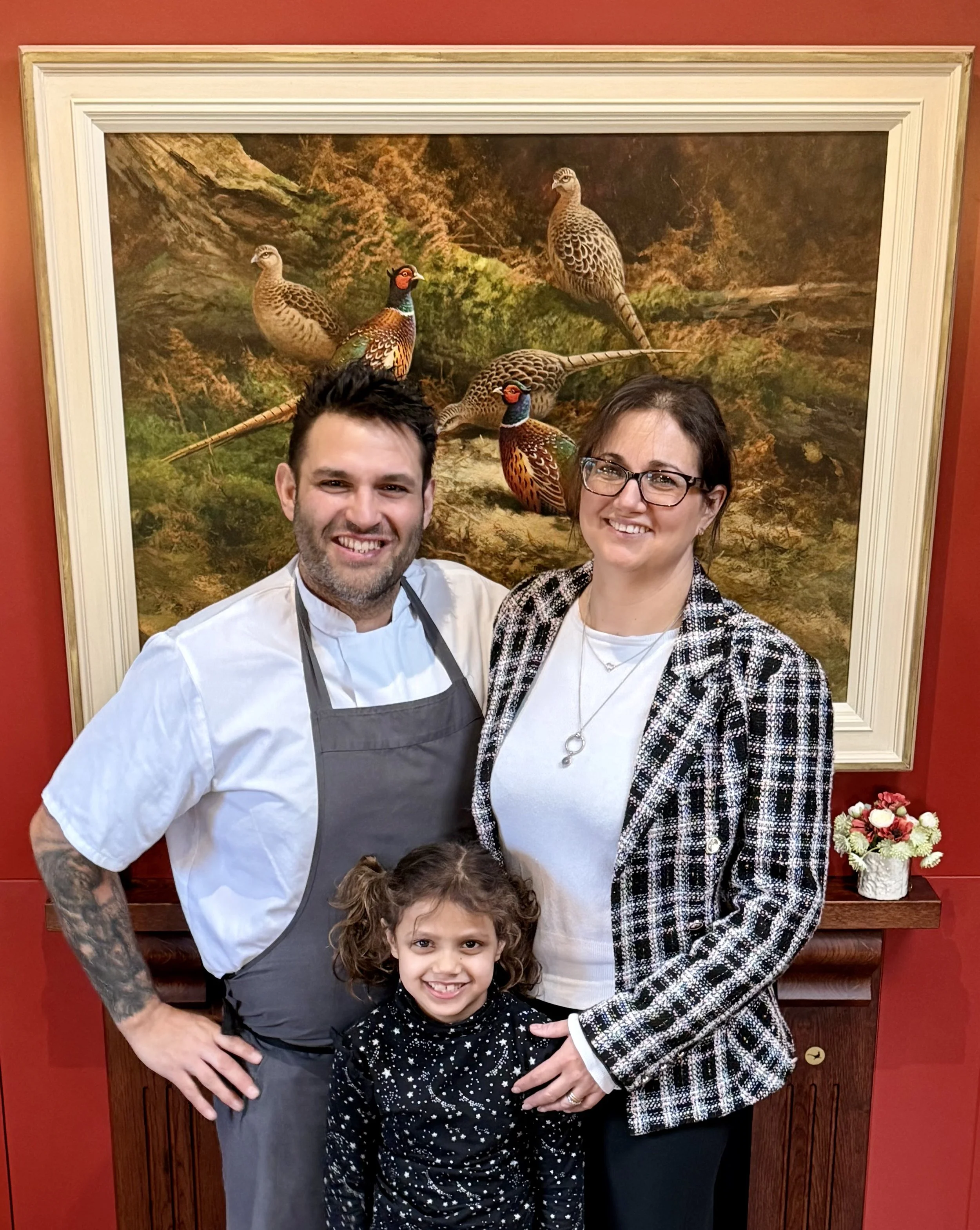 A family of three smiling and posing in front of a framed painting of pheasants, with a man wearing a white shirt and gray apron, a woman with glasses in a black and white checkered blazer, and a young girl in a black star-patterned top.