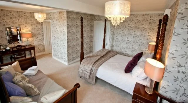 A bedroom with floral wallpaper, a four-poster bed with white bedding and purple pillows, a wooden nightstand with a lamp, and a chandelier hanging from the ceiling.