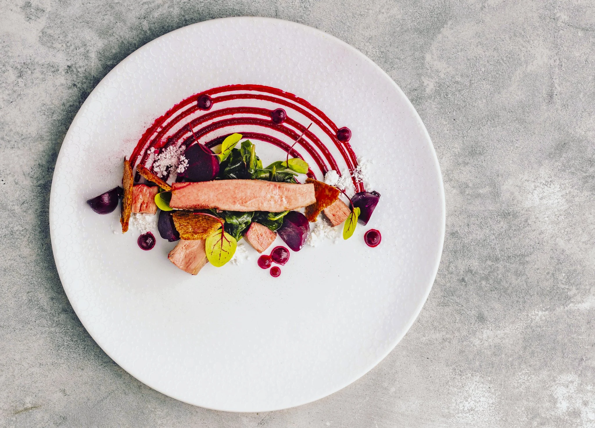 Artistic plate of pink cooked chicken garnished with cherry tomatoes, green leaves, berry sauces, and edible flowers, presented with a red swirl sauce on a white textured plate.