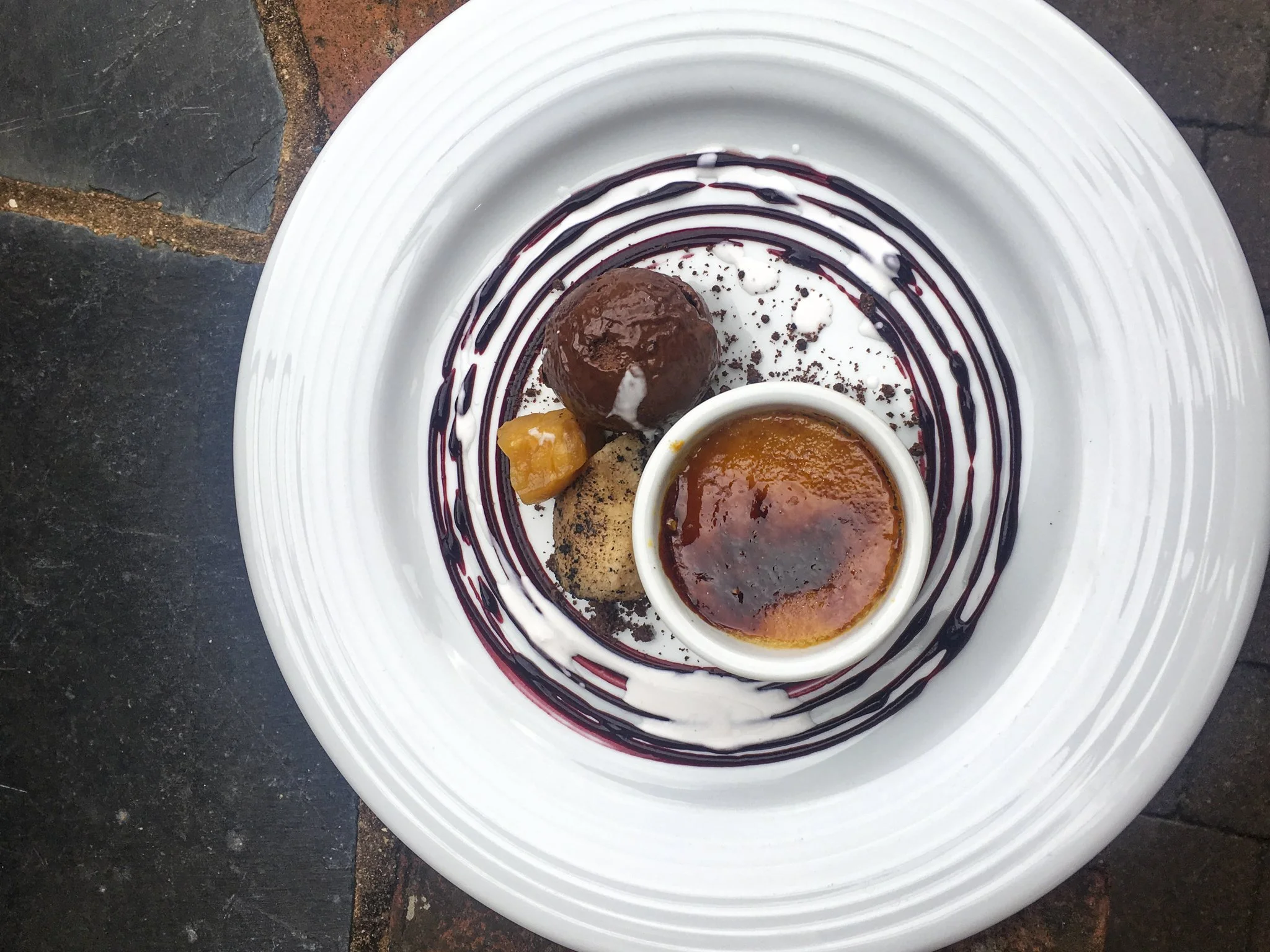 A dessert plate with a small ramekin of caramel flan, a chocolate truffle, a cookie, and a piece of fruit, decorated with chocolate and white sauces.