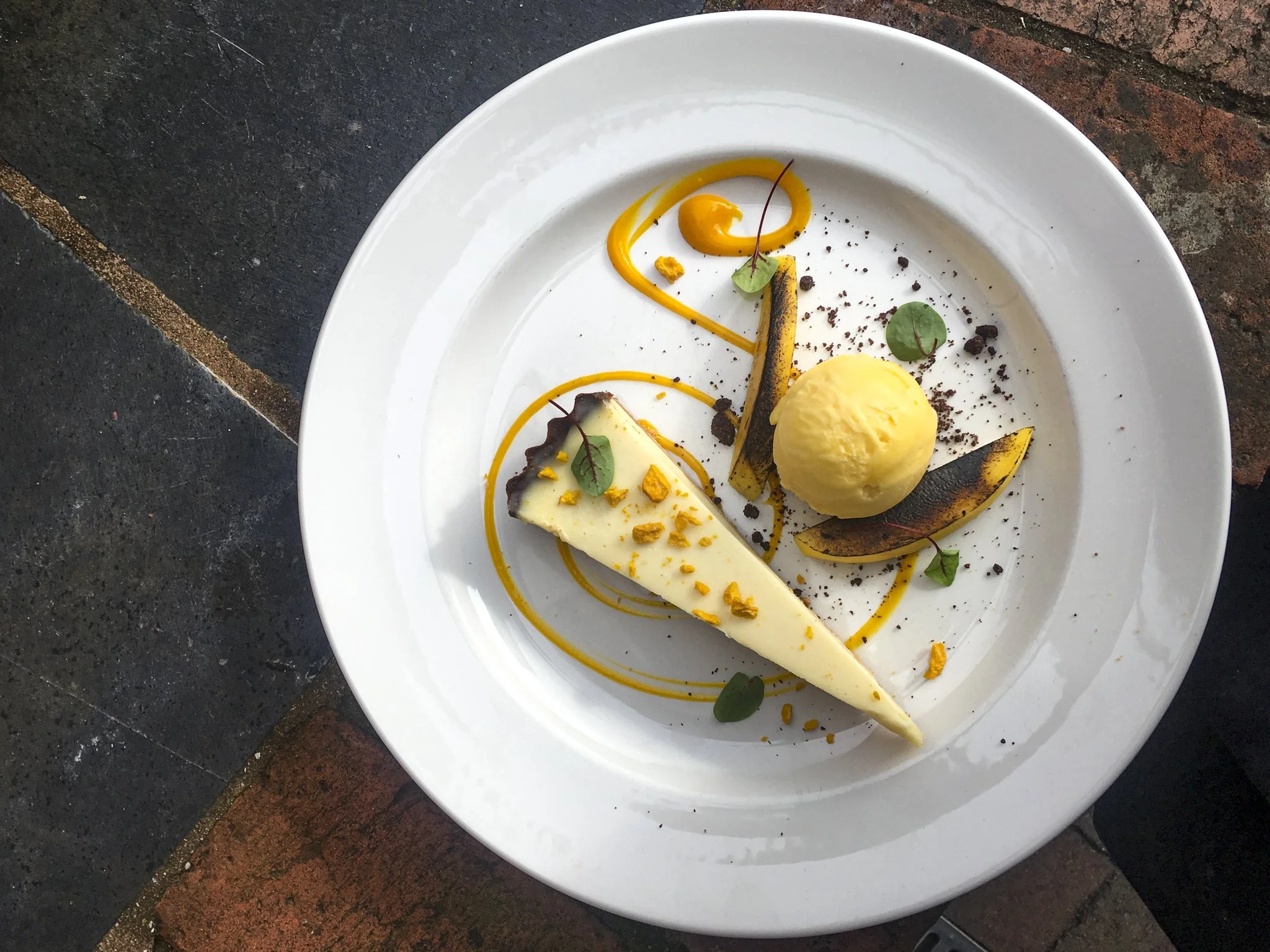 Empty white plate with a slice of cheesecake, scoop of vanilla ice cream, grilled fruit slices, and garnishes, on a dark and brick surface.