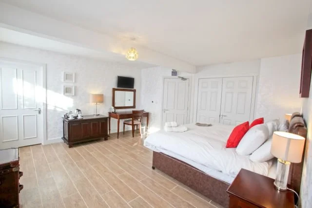 Hotel room with a large bed, nightstand, TV, dresser, and doors, decorated in white and wood tones.