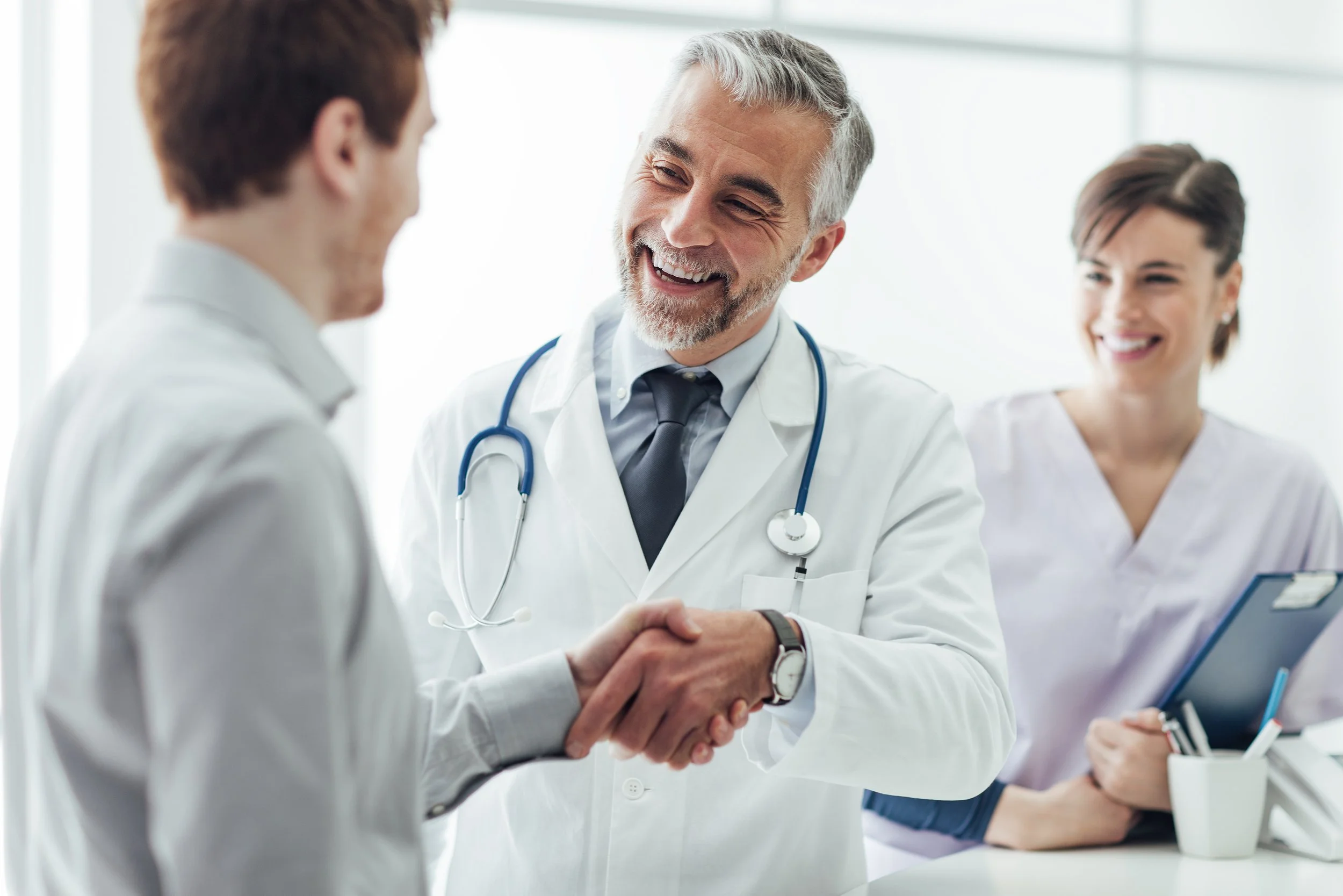 A happy male doctor with gray hair and beard shaking hands with a patient in a medical office, with a female nurse smiling in the background.