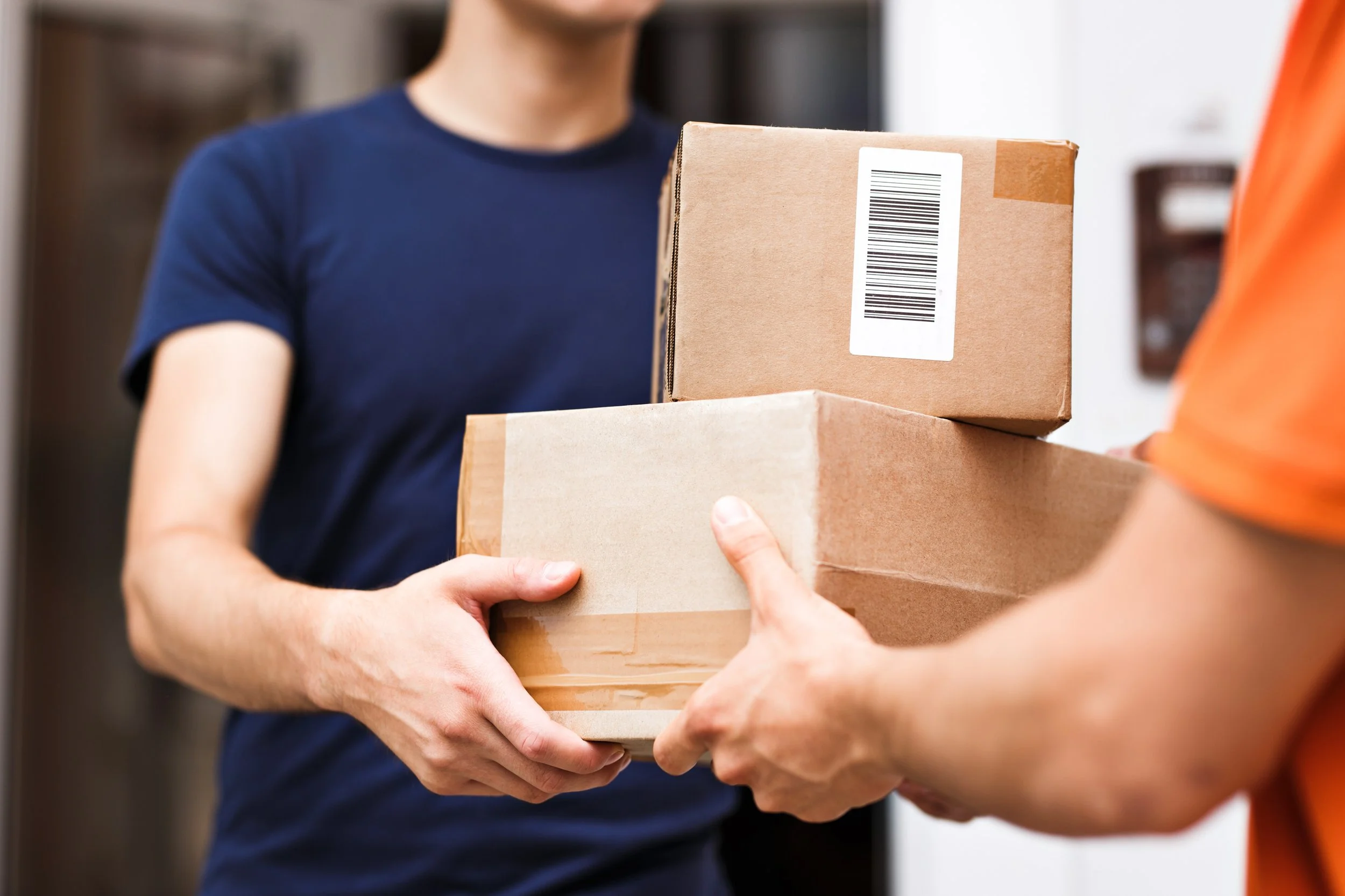 Person wearing a blue shirt handing over two cardboard boxes to another person in an orange shirt.