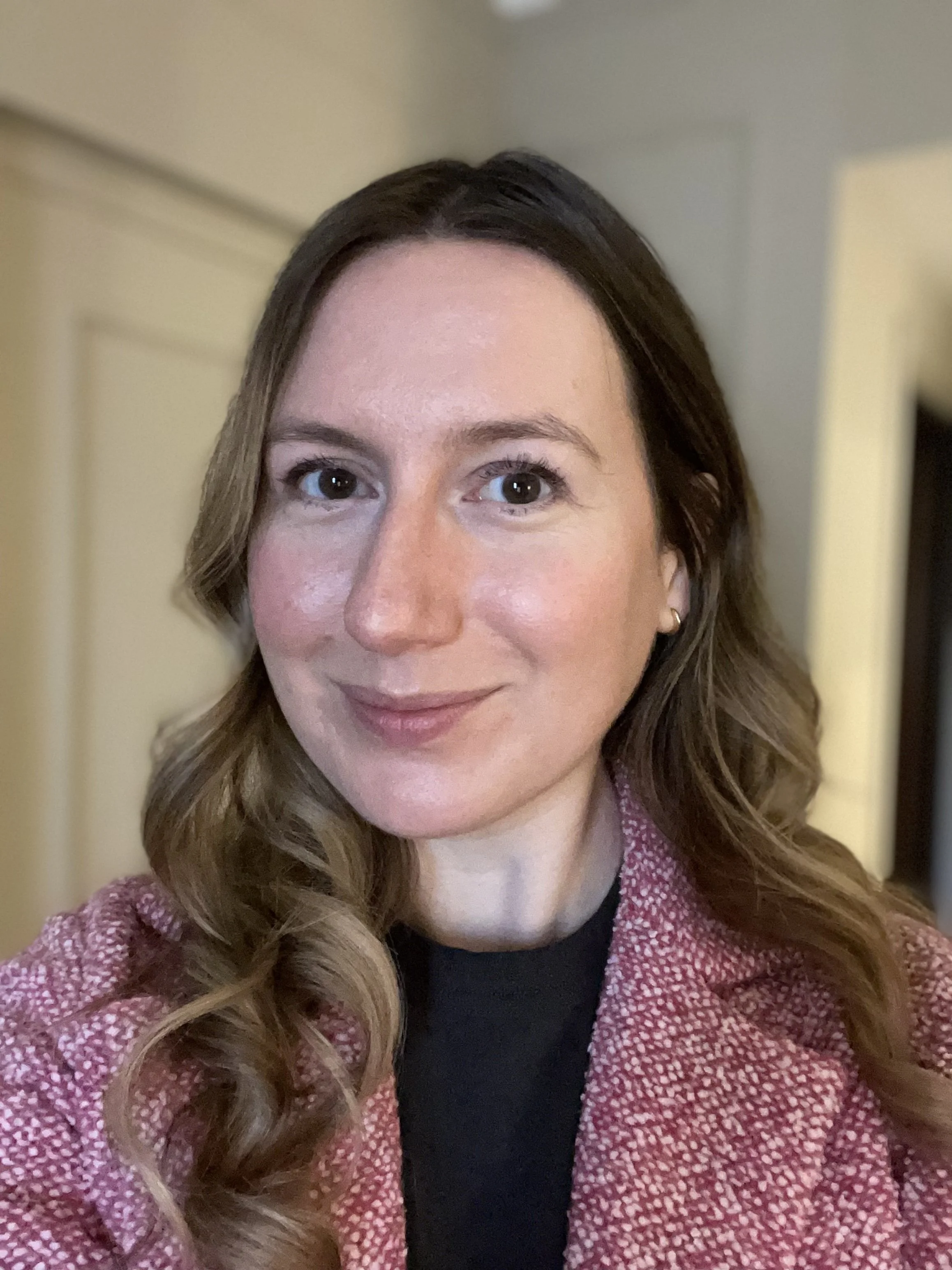 A woman with brown hair and fair skin smiling at the camera, wearing a pink patterned blazer and a black top, with a blurred indoor background.