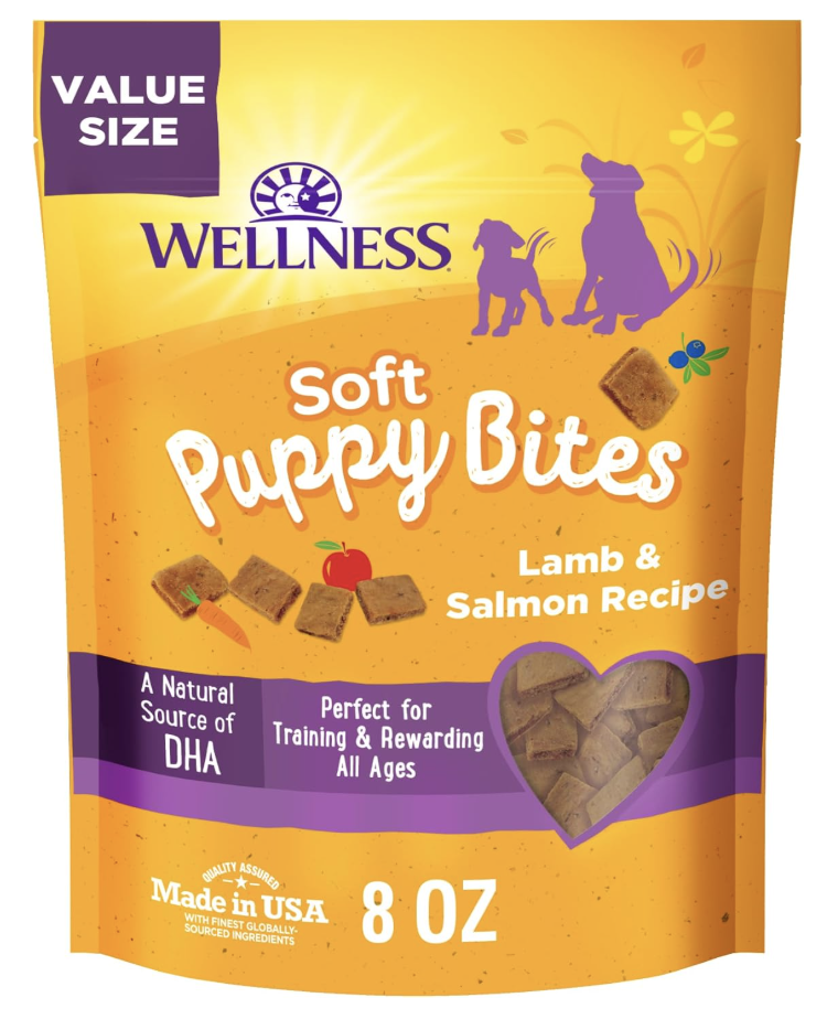 Packaging of Wellness Soft Puppy Bites dog treats in lamb and salmon flavor, 8 ounces, with a purple and orange design, silhouettes of a dog and puppy in the background, and a heart-shaped window showing the treats inside.
