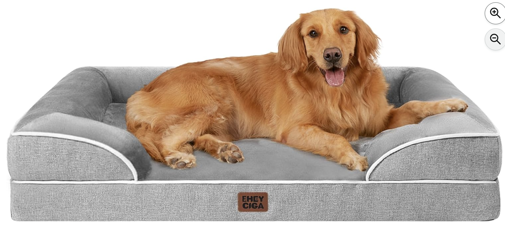 Golden retriever lying on a gray dog bed with white piping, looking at the camera with its tongue out.
