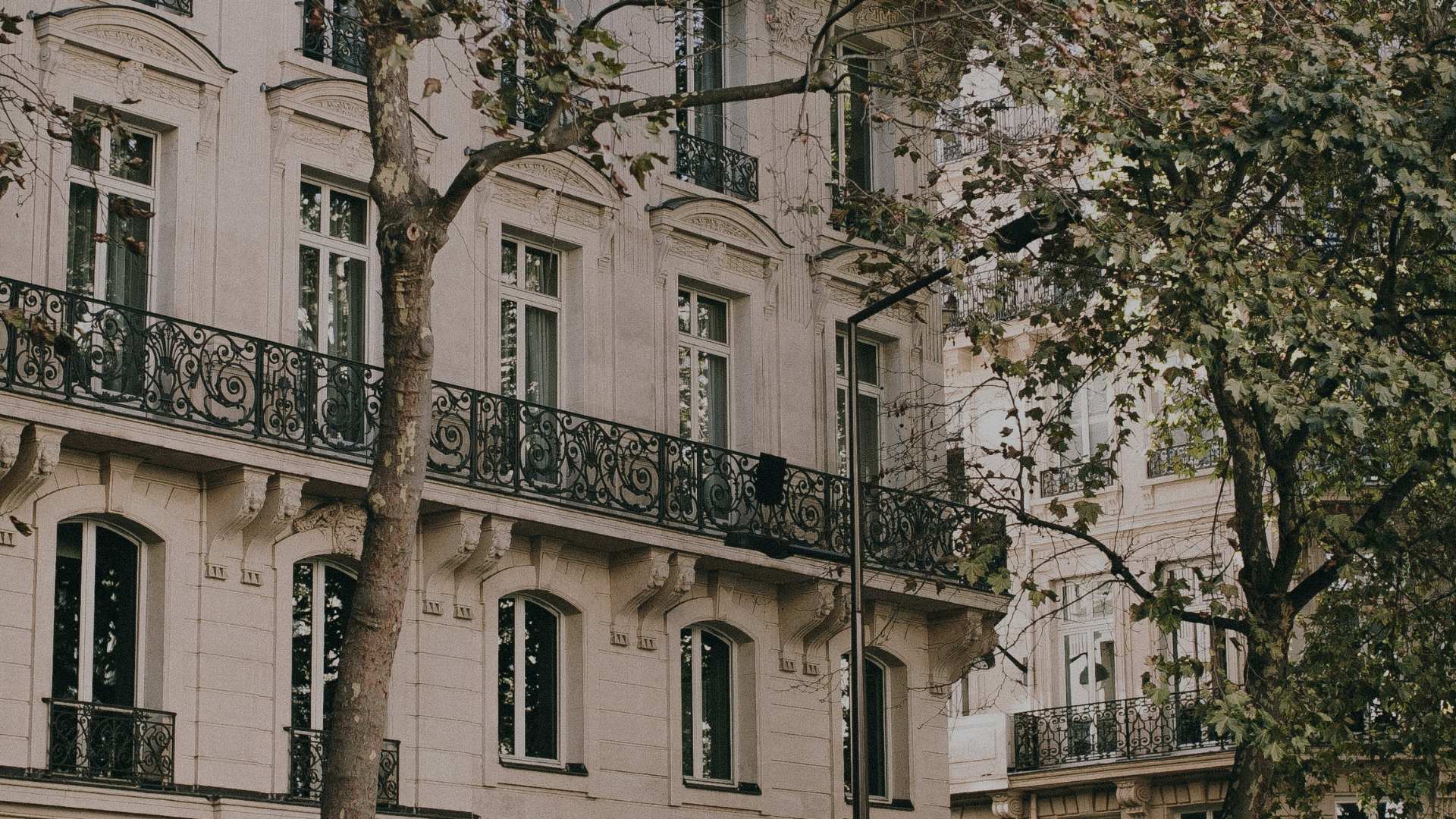 Photo of a classic European building with ornate stone facade, tall windows, and wrought iron balconies, partially obscured by trees with green leaves.