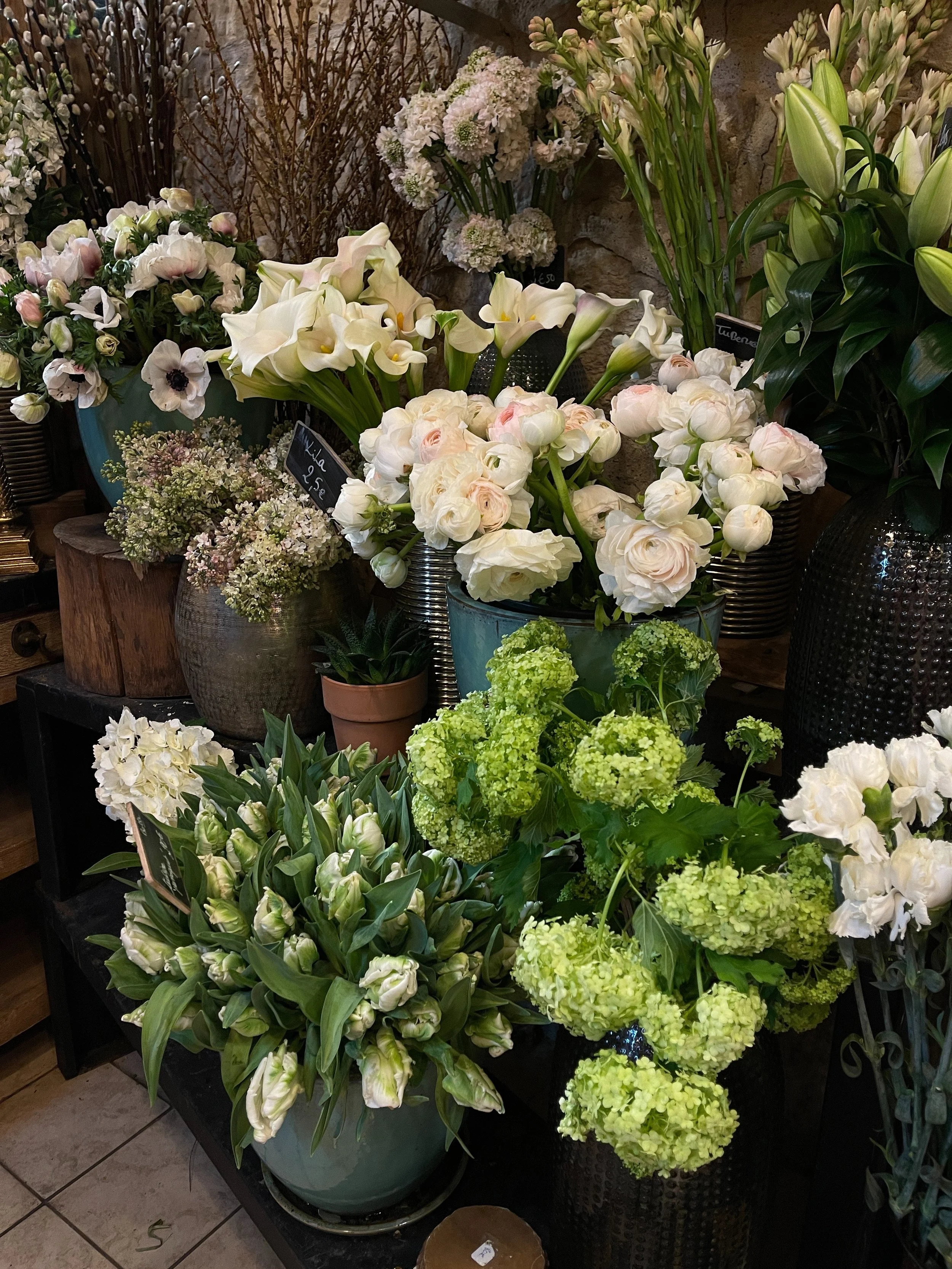 Various white and pale pink flowers including calla lilies, lilies, and hydrangeas arranged in pots and vases in a flower shop.