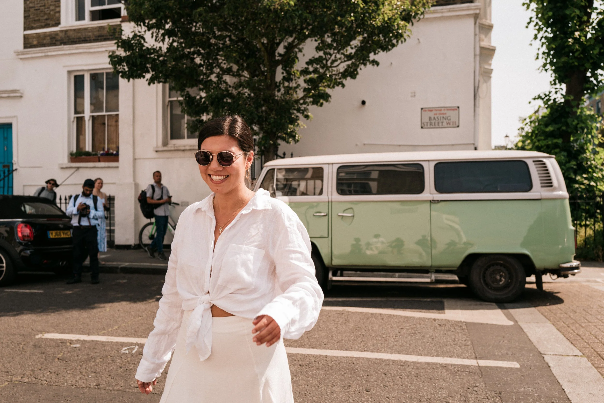 A woman wearing sunglasses and a white shirt smiling, walking in a parking lot with a vintage green and white van behind her and a white building with trees in the background.