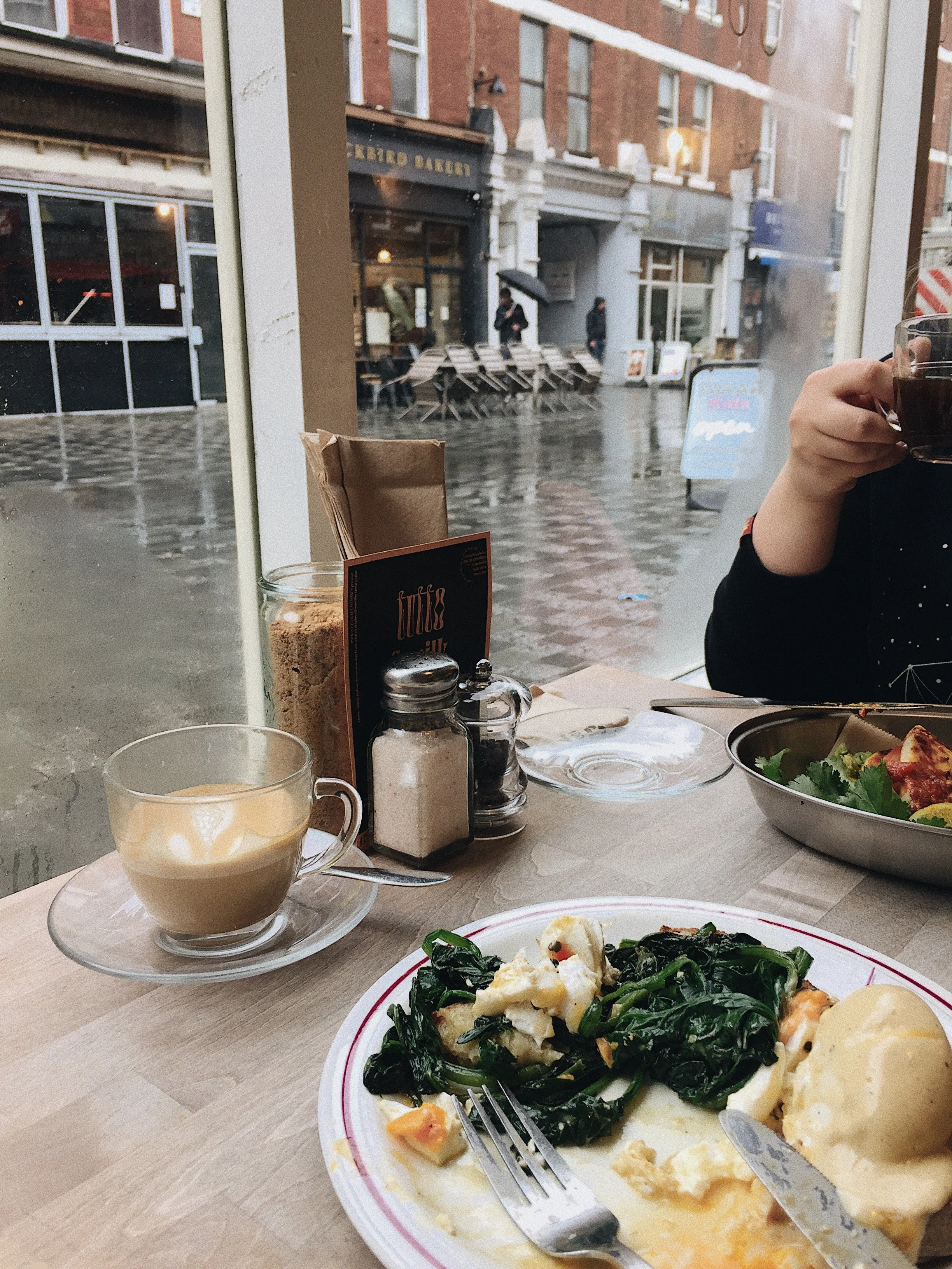View through a large restaurant window showing a rainy urban street scene with pedestrians. Inside, a table with a plate of spinach and cheese, a coffee drink, and condiments.