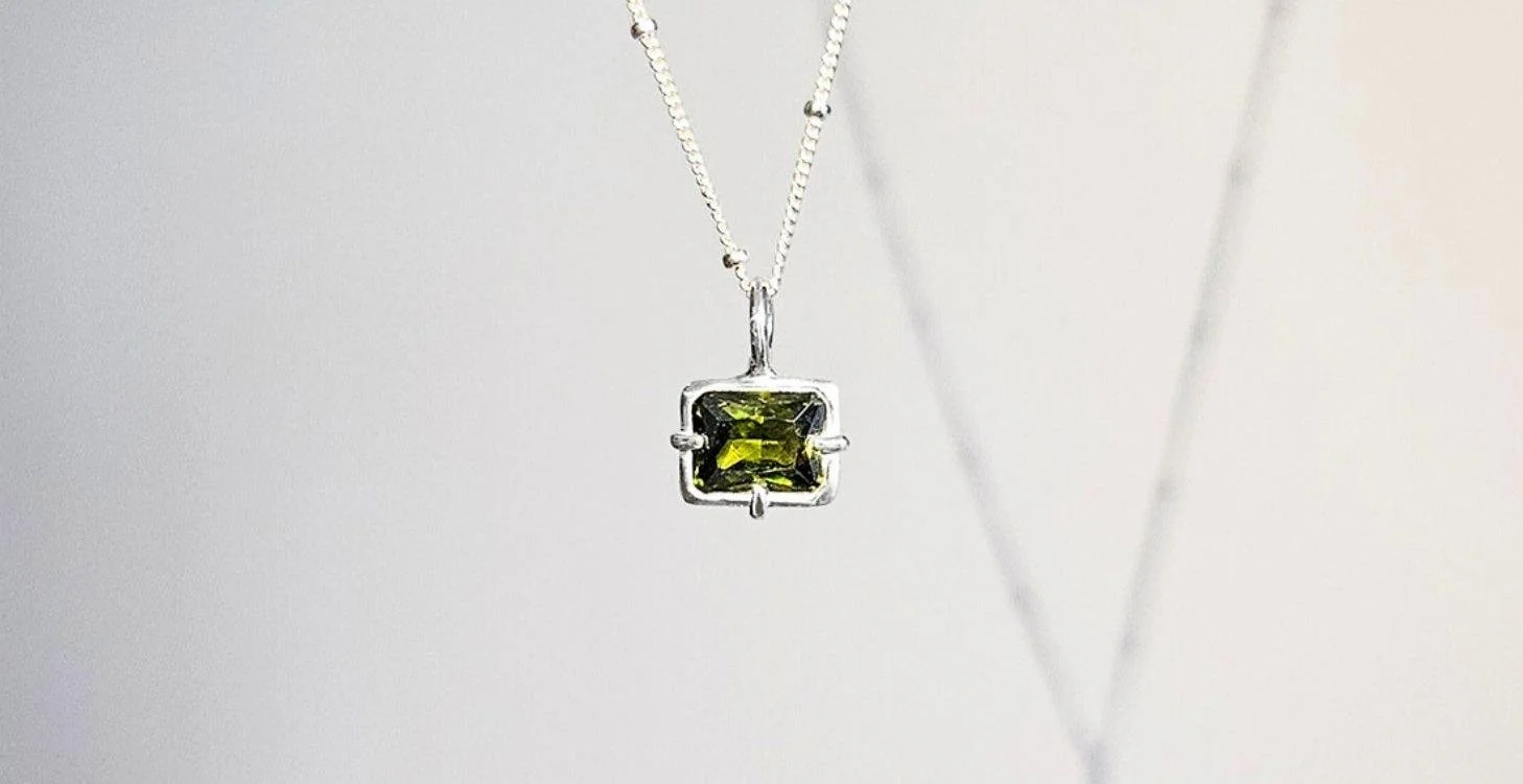 A silver necklace with a square-shaped green gemstone pendant hanging against a plain, light-colored background.