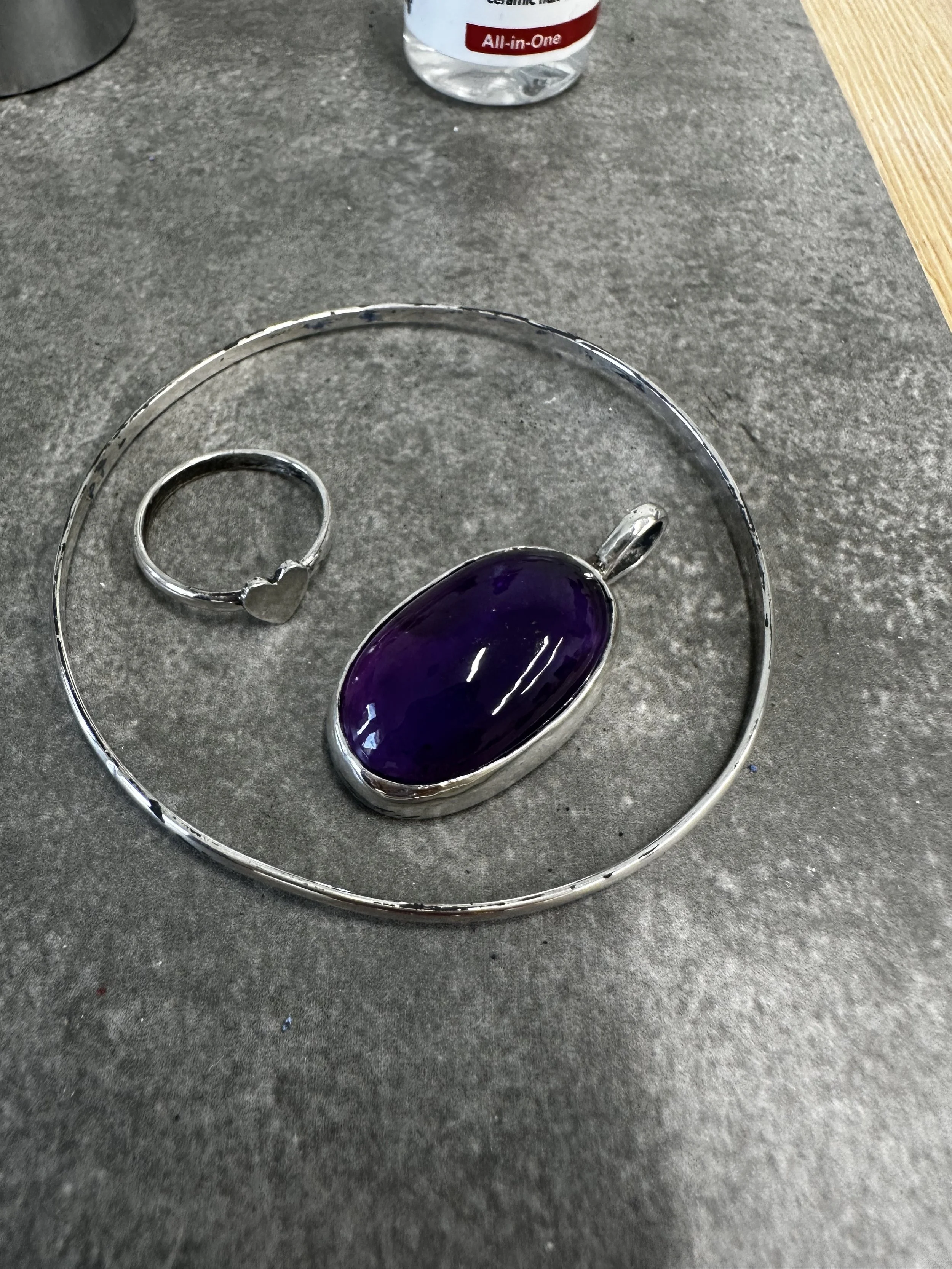 Close-up of a silver jewelry tray with an oval-shaped purple stone ring and a silver heart-shaped ring on a gray surface with a bottle in the background.