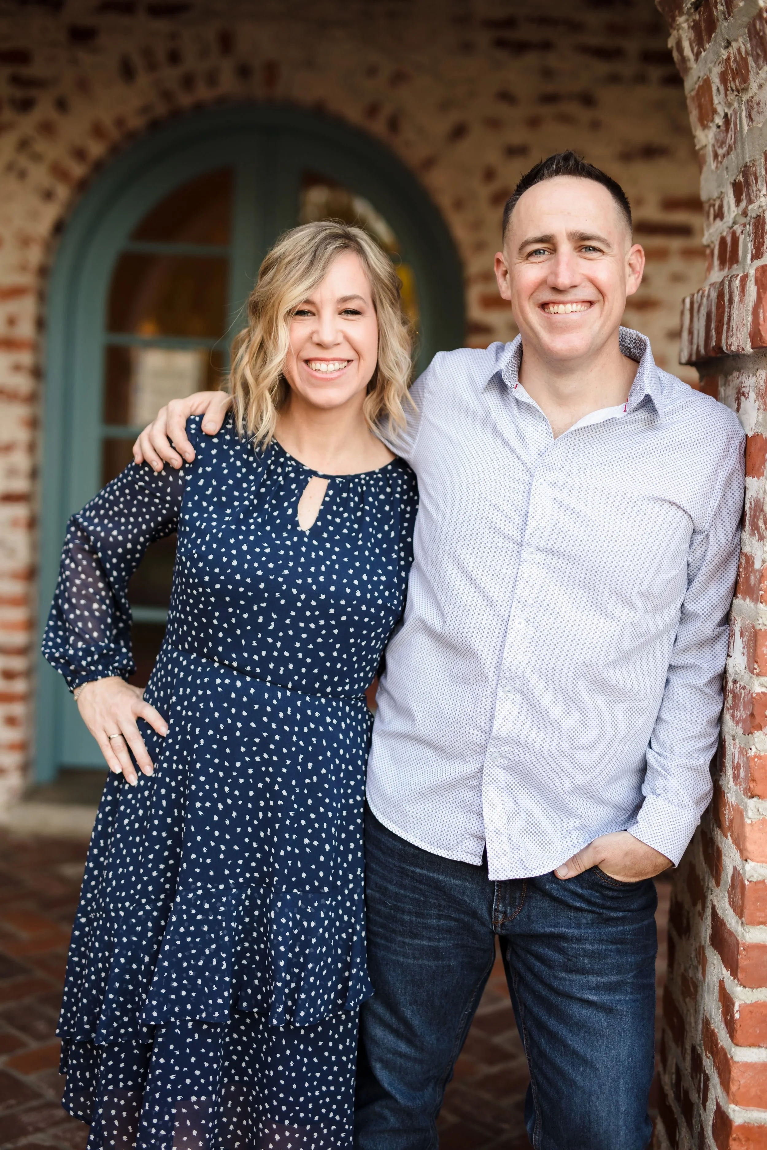 Smiling man and woman standing close together outside, with a brick wall and an arched window in the background. The woman has blonde wavy hair and is wearing a navy blue dress with white polka dots. The man has short dark hair, is wearing a light-colored button-down shirt and jeans, and has his arm around the woman.